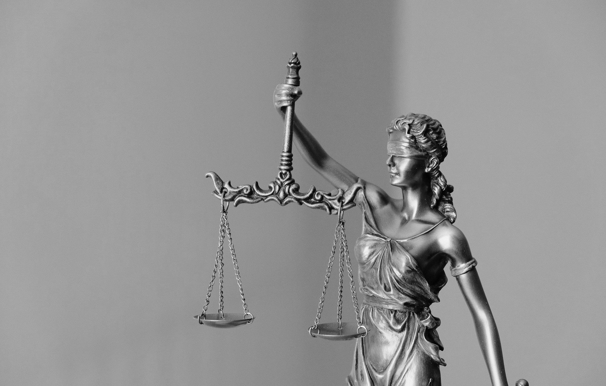 L'image représente une statue allégorique de la justice. La figure féminine, vêtue d'une robe drapée, tient une balance dans une main, symbole d'équité et de mesure. Elle porte un bandeau sur les yeux, incarnant l'impartialité et l'objectivité. La statue est en métal et présente des détails raffinés, tels que des mèches de cheveux tressées. Le fond est simple et neutre, mettant en valeur la silhouette de la figure tout en accentuant son importance symbolique.