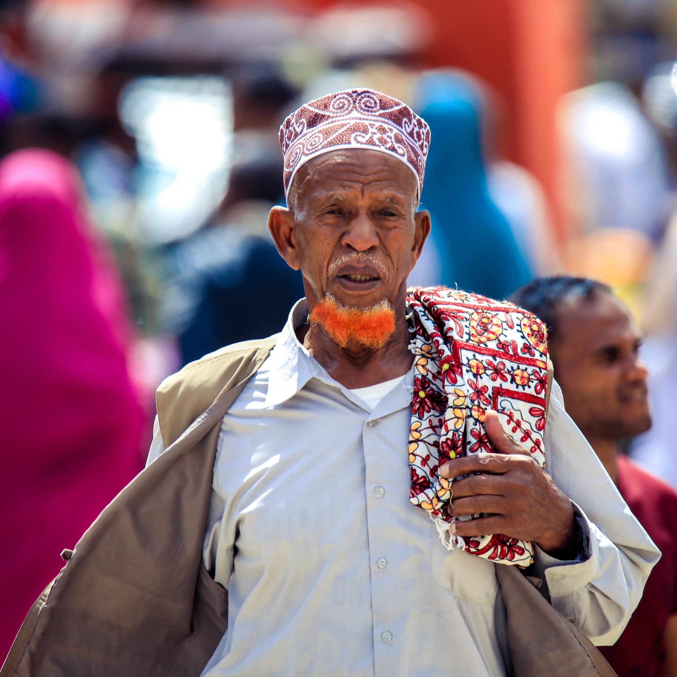 L'image montre un homme d'un certain âge marchant dans une rue animée. Il porte une tenue traditionnelle, avec une chemise claire et un grand châle coloré drapé sur son épaule. Sa tête est couverte d'un bonnet à motifs distinctifs, et il a une barbe teintée en orange, qui attire l'attention. En arrière-plan, on aperçoit des silhouettes floues de personnes vêtues de vêtements colorés, créant une ambiance vivante et dynamique. Les couleurs et la diversité des tenues témoignent d'un environnement culturel riche.