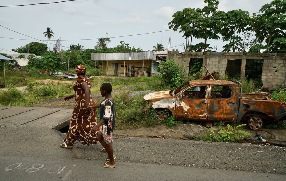 L'image montre une scène d'une rue dans un environnement rural ou semi-urbain. Sur le trottoir, une femme vêtue d'une longue robe ornée de motifs blancs marche aux côtés d'un jeune garçon. Ils avancent côte à côte, probablement en direction d'un endroit précis. En arrière-plan, on peut voir un paysage verdoyant avec des arbres, ainsi que des bâtiments partiellement construits, évoquant une ambiance de quartier en développement. À droite, un vieux véhicule, rouillé et en piteux état, est abandonné sur le côté de la route, entouré de végétation. Le ciel est nuageux, suggérant qu'il pourrait faire chaud et humide. L'ensemble dégage une atmosphère à la fois paisible et un peu nostalgique.