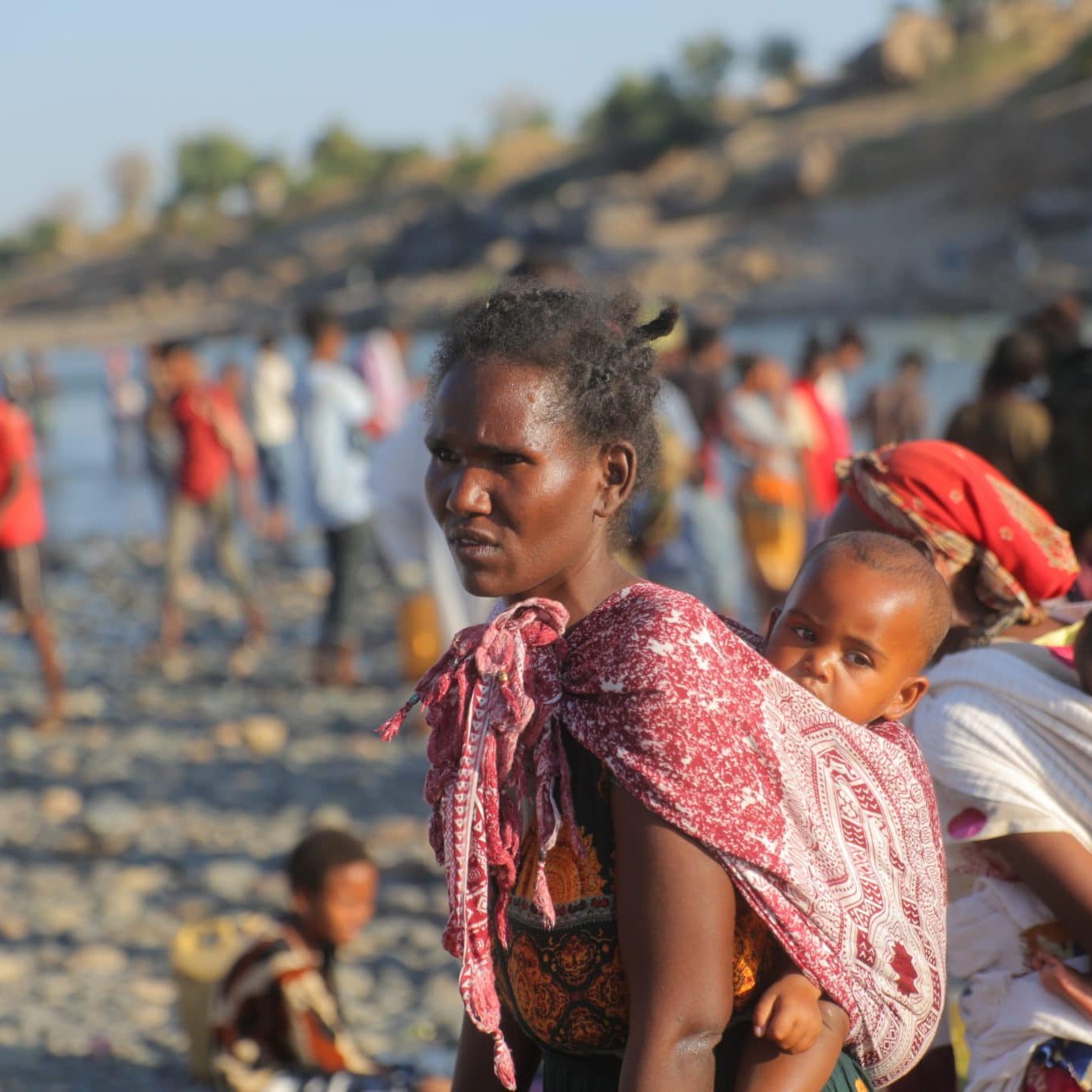 L'image représente une scène vibrante et émotive d'un groupe de personnes au bord d'une rivière. Au premier plan, une femme porte un enfant sur son dos, enveloppé dans un tissu coloré. Son expression est sérieuse et déterminée. Elle est entourée d'autres personnes, dont certaines portent également des enfants. Les vêtements des personnes sont variés, avec des motifs et des couleurs vives. En arrière-plan, on aperçoit des rochers et une végétation légère, ainsi que d'autres silhouettes de personnes se déplaçant le long de la rive. L'atmosphère semble chargée d'une mixité d'activités et d'émotions, reflétant à la fois la vie quotidienne et des moments de transition.