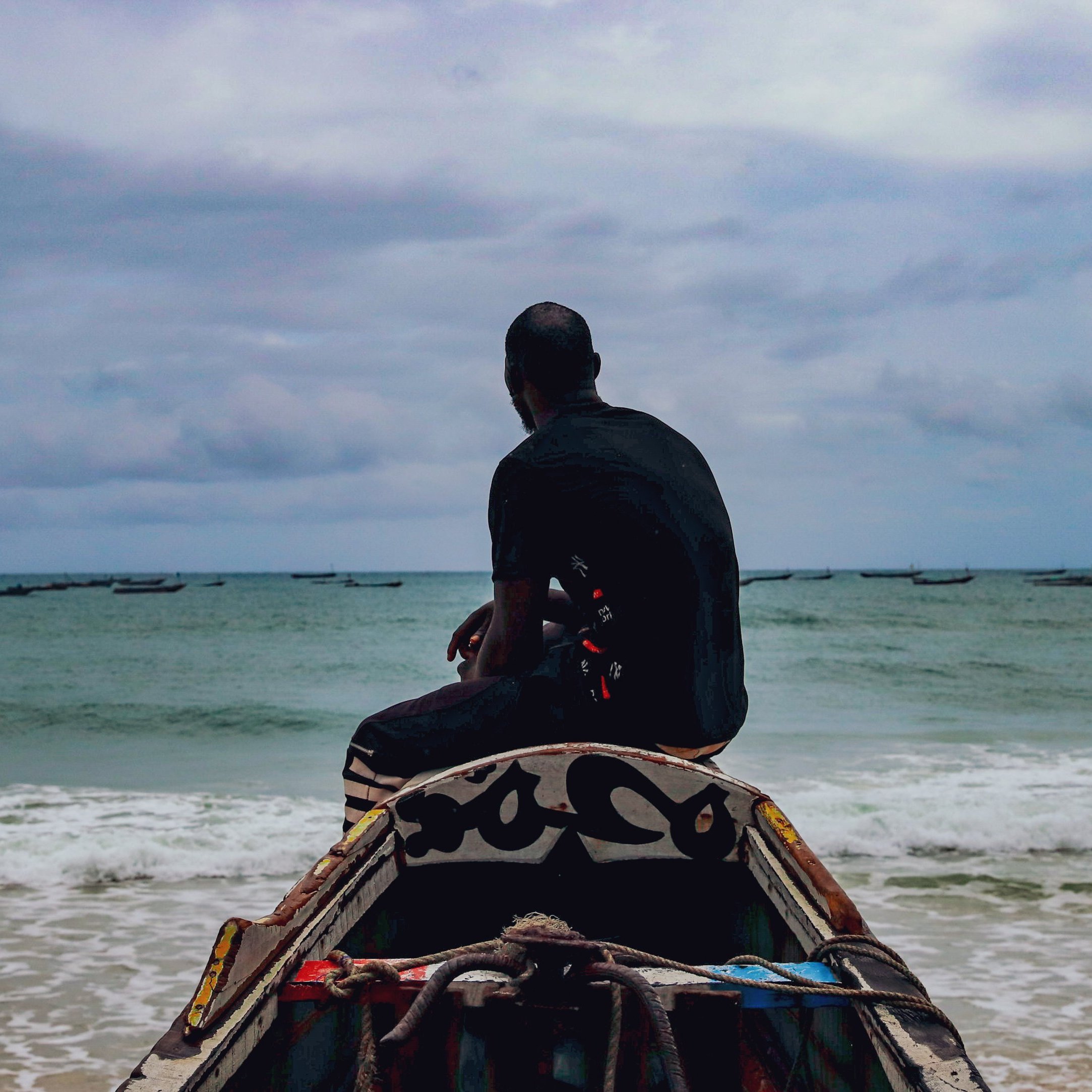 L'image montre un homme assis sur une barque, tourné vers la mer. Il regarde l'horizon, où l'eau rencontre le ciel nuageux. La mer est calme avec quelques petites vagues, et on aperçoit des bateaux au loin. L'ambiance est sereine, soulignée par le ciel gris et la lumière douce. L'homme porte un vêtement sombre et semble pensif, contemplant la vastitude de l'océan devant lui.