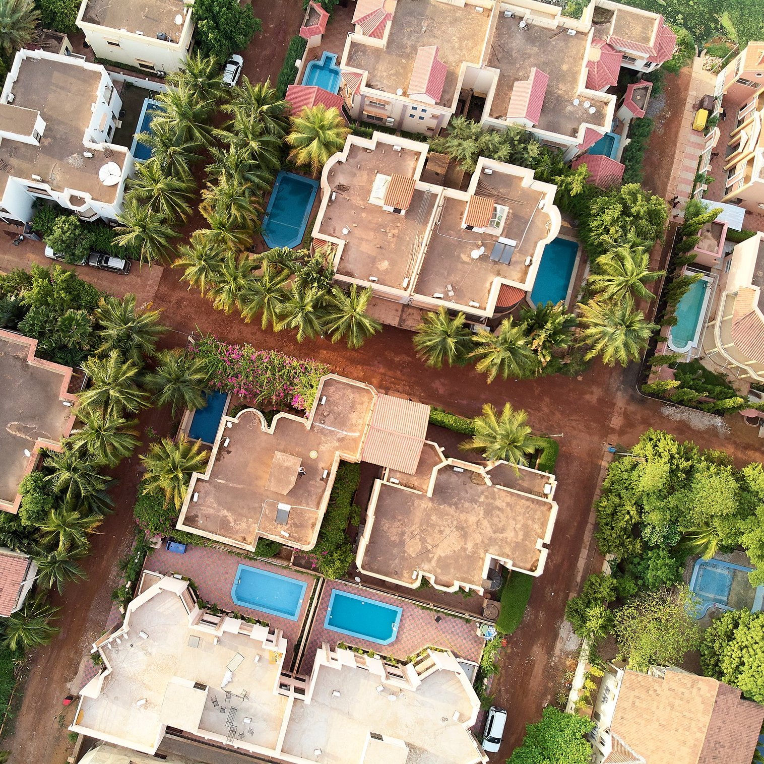 L'image montre une vue aérienne d'un complexe résidentiel. On y voit plusieurs maisons aux toits plats, disposées autour de petites piscines bleues. Parmi ces maisons, il y a de nombreuses palmiers qui ajoutent une touche de verdure. Les allées sont en terre et les maisons sont agencées de manière à créer un réseau urbain harmonieux. Les couleurs de la terre et des toits se marient pour donner une ambiance chaleureuse et accueillante au paysage.