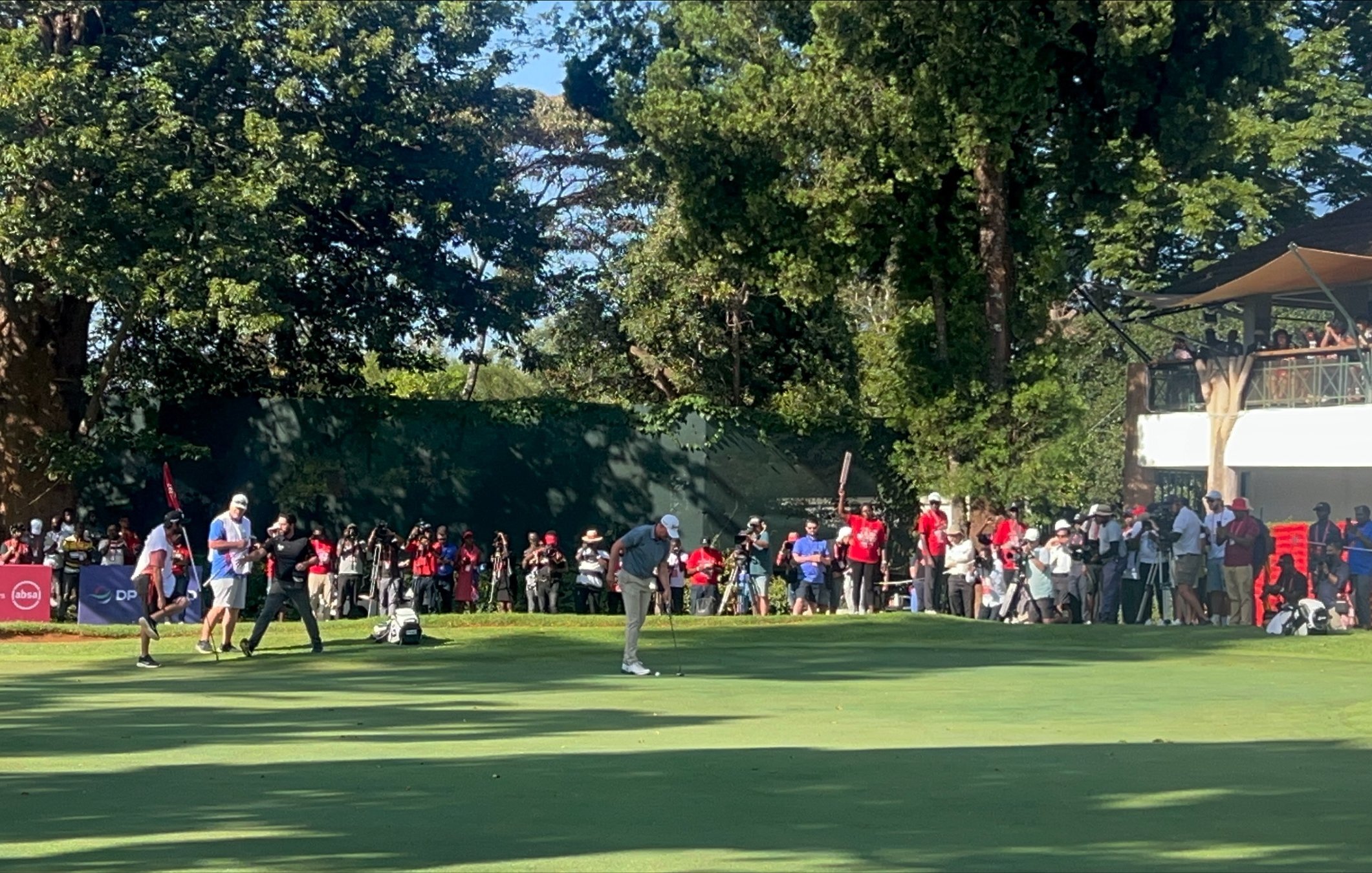 Imaginez un vaste terrain de golf, baigné par la lumière du soleil. Au premier plan, deux joueurs se concentrent sur le putting green, faisant preuve de délicatesse et de précision. L'un d'eux est en position de putt, tandis que l'autre observe attentivement. À l'arrière-plan, des spectateurs en tenue décontractée, vêtus de couleurs variées, attendent avec excitation, certains filmant ou prenant des photos. Autour du green, de grands arbres fournissent de l'ombre, ajoutant à la beauté de l'environnement. À droite, on aperçoit un bâtiment avec des balcons, où d'autres personnes sont rassemblées pour profiter du spectacle. L'atmosphère est vivante, remplie de murmures, de rires et de l'anticipation d'un moment décisif dans le jeu.