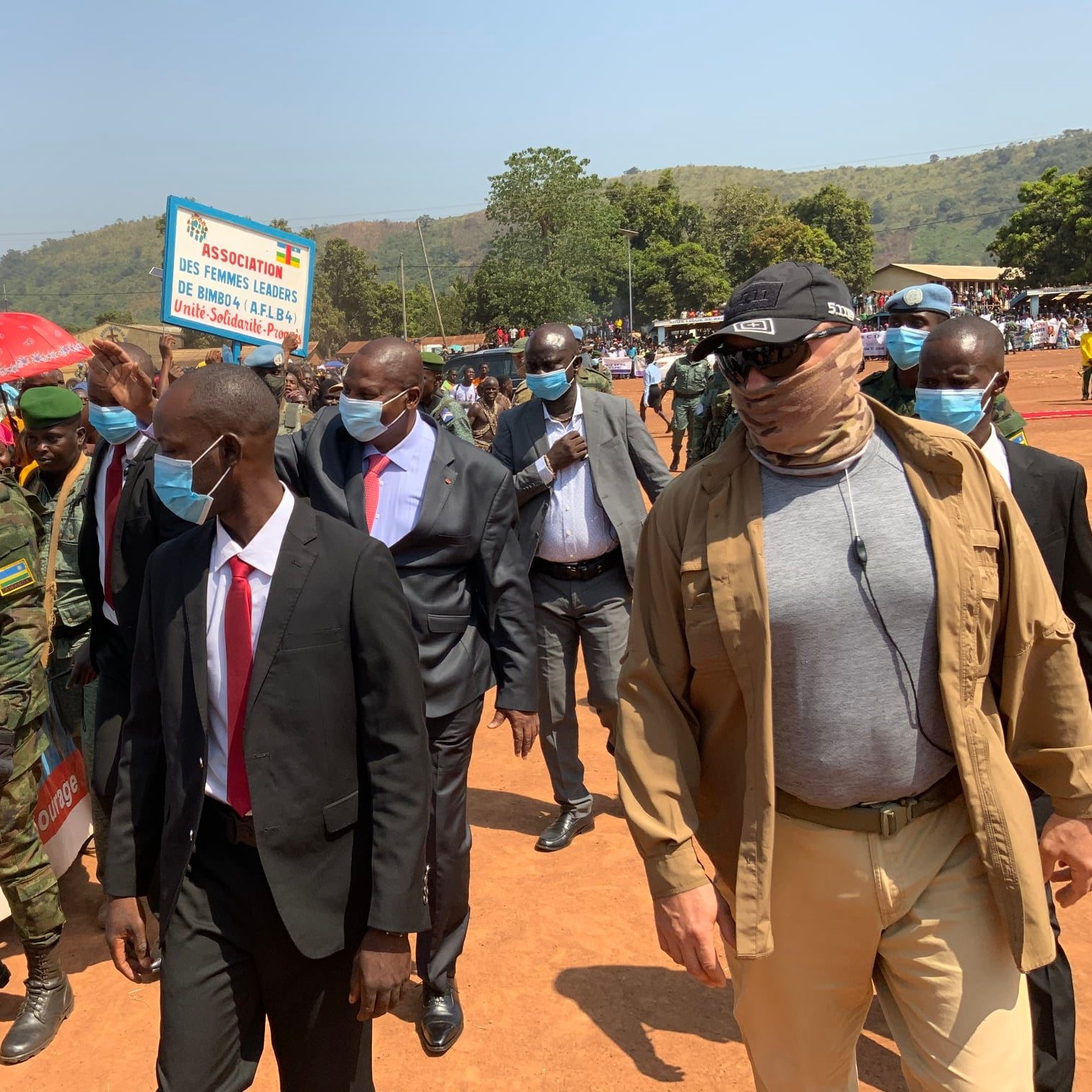Dans cette image, un groupe de personnes est rassemblé lors d'un événement en extérieur. Au premier plan, on voit plusieurs hommes en costume, parmi lesquels un homme en particulier porte des lunettes de soleil et un foulard. Ils avancent au milieu d'une foule. À gauche, des militaires en uniforme surveillent la situation. En arrière-plan, des gens brandissent des pancartes, et des parasols colorés ajoutent une ambiance vivante à la scène. Le décor est verdoyant, avec des collines en arrière-plan sous un ciel clair. L'atmosphère semble festive et dynamique.