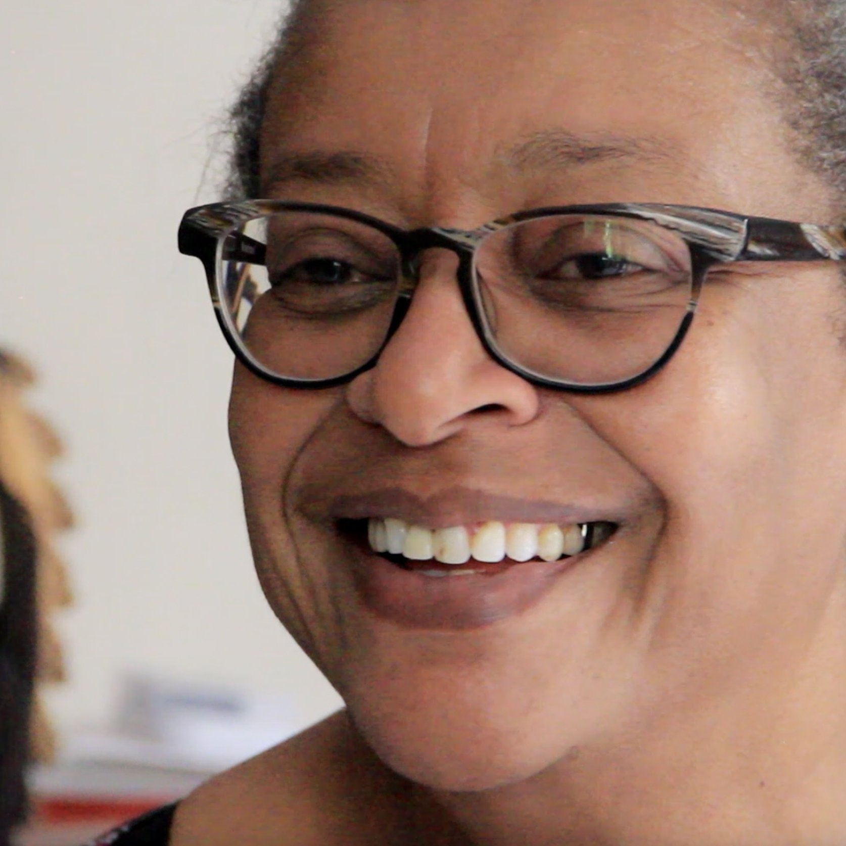 L'image montre une femme souriante, avec des lunettes à monture noire. Son expression est chaleureuse et accueillante. Elle a les cheveux grisonnants, peignés en arrière, et porte des boucles d'oreilles discrètes. En arrière-plan, on aperçoit une sculpture en bois, qui semble représenter un motif stylisé. L'atmosphère est conviviale et intime, suggérant un moment de bonheur ou de partage.