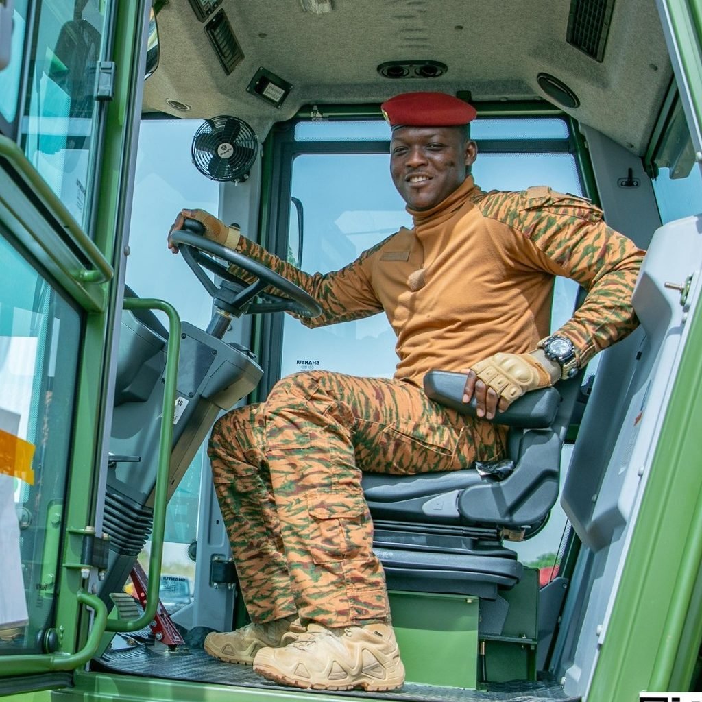 Dans cette image, nous voyons un homme assis à l'intérieur d'un tracteur. Il porte un uniforme couleur sable orné de motifs camouflés et un béret rouge. Son expression est confiante et il sourit, montrant un sentiment de fierté. À l'arrière-plan, on aperçoit d'autres personnes habillées de manière similaire, travaillant dans un environnement agricole. Le ciel est clair et lumineux, suggérant une journée ensoleillée. L'ensemble de la scène dégage une atmosphère de travail collectif et de détermination.