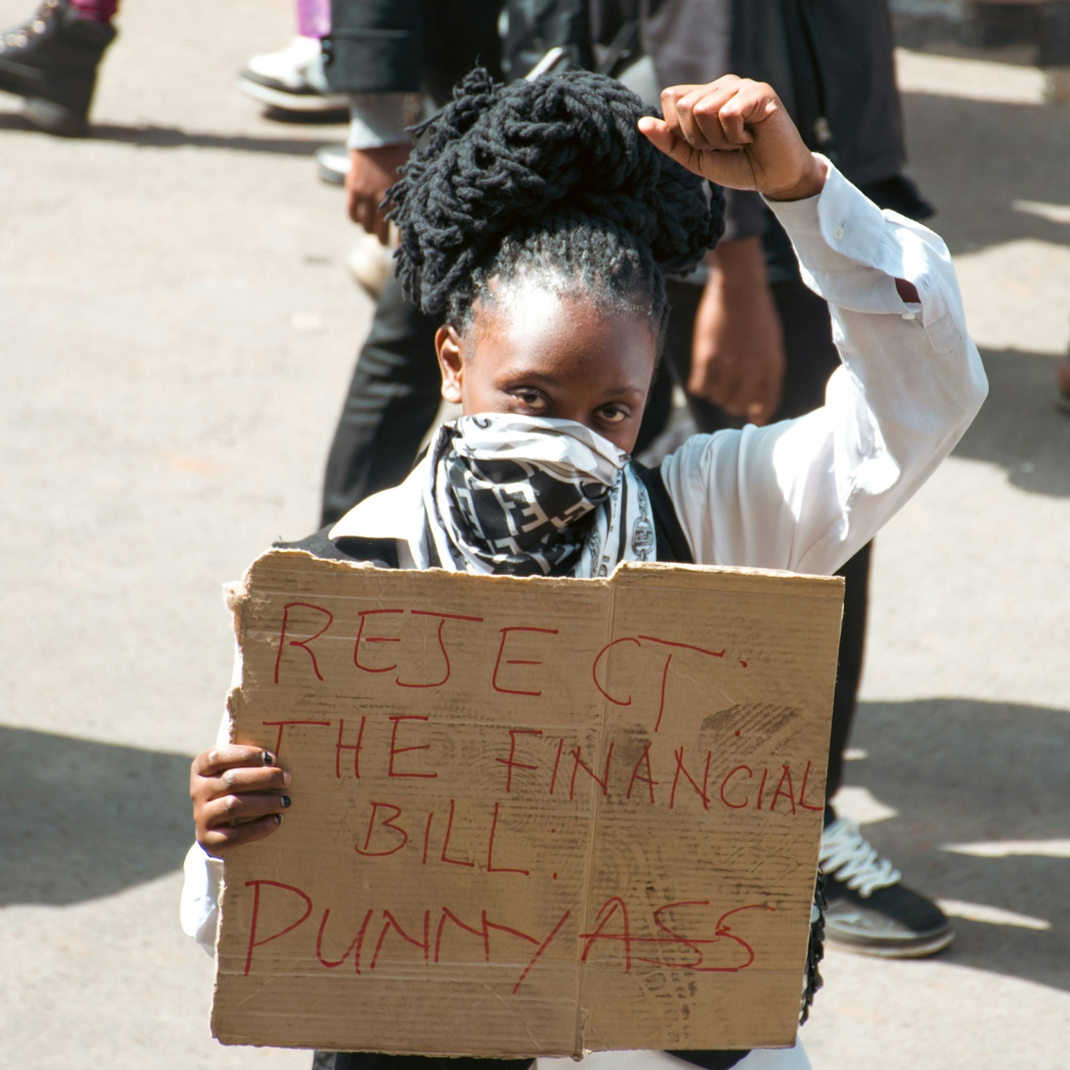 L'image montre une manifestation dans laquelle une femme se tient au premier plan. Elle a des cheveux noirs et crépus coiffés en chignon. Elle porte un masque sur le visage et un haut blanc. Dans sa main, elle élève le poing, symbole de résistance. L'autre main tient une pancarte en carton sur laquelle sont inscrits en lettres rouges les mots "REJECT THE FINANCIAL BILL PUNNYASS". En arrière-plan, on distingue d'autres personnes, soulignant le caractère collectif de la protestation. L'atmosphère est chargée d'énergie, avec un sentiment d'engagement palpable.