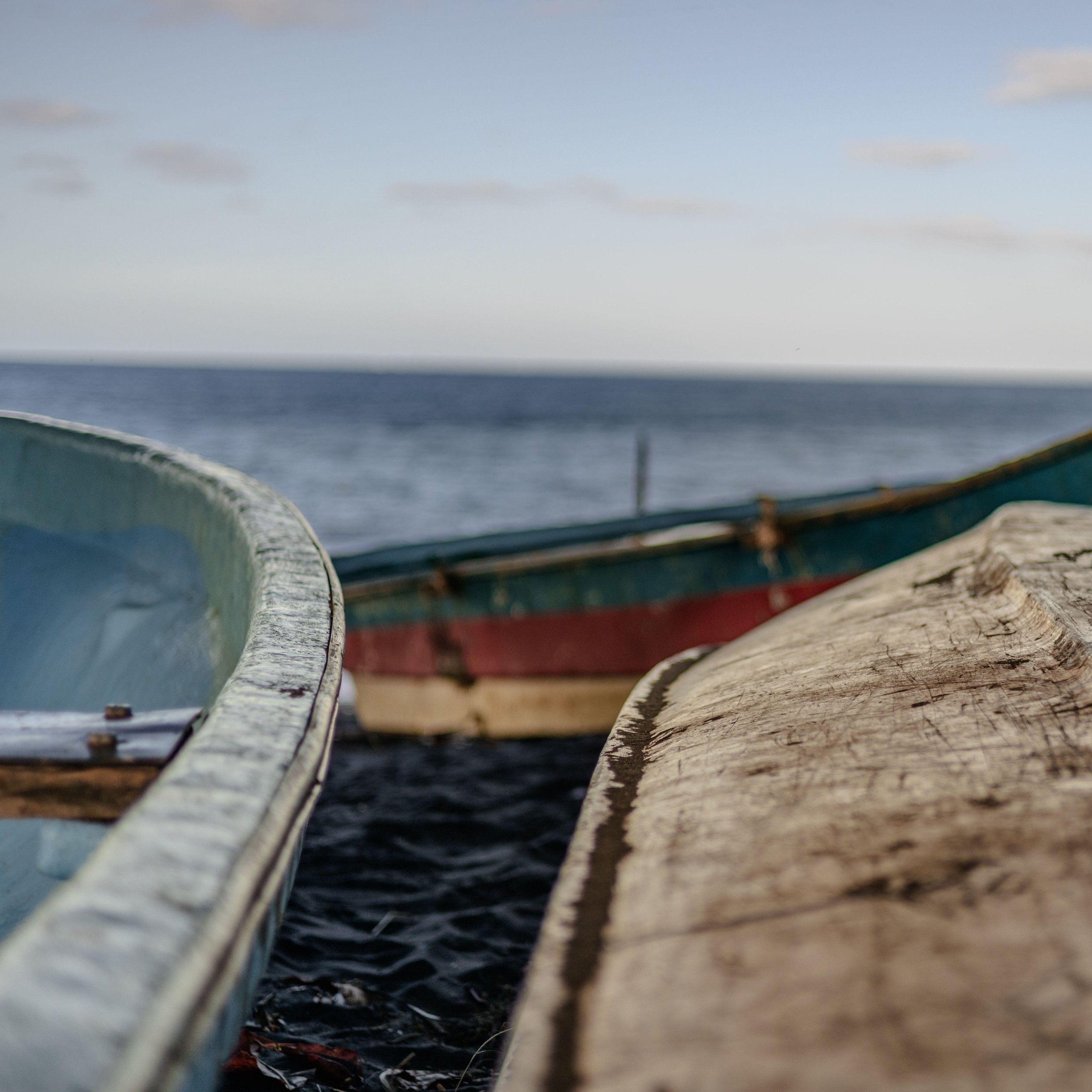 Dans cette image, vous pouvez imaginer un décor paisible au bord de l'eau. Deux bateaux en bois, de designs simples, sont amarrés près d'une plage. Le premier bateau a une structure légèrement arrondie avec une peinture bleu clair sur les côtés, tandis que le second, plus allongé, présente des nuances de bleu et de rouge. Les bateaux sont usés, témoignant de leur utilisation fréquente. En arrière-plan, l'eau s'étend à l'horizon, calme et réfléchissante, et le ciel est dégagé avec quelques nuages légers. L'ambiance est tranquille et maritime, invitant à la contemplation.