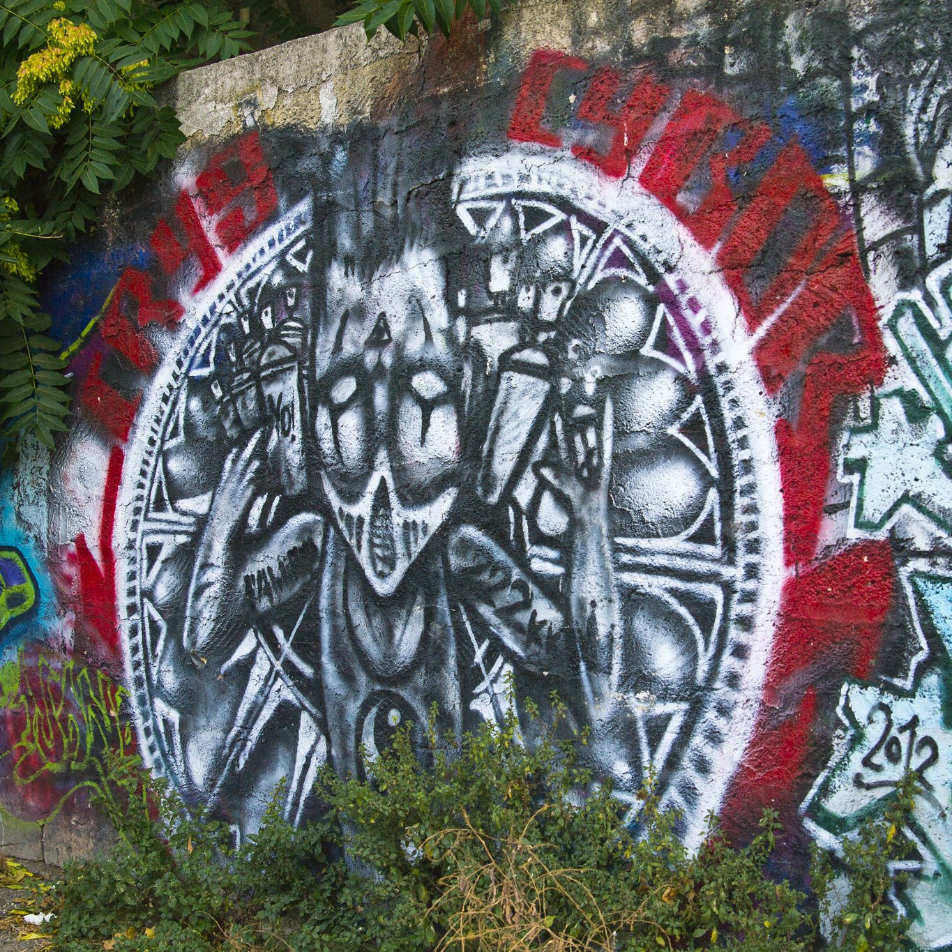 L'image représente une peinture murale vibrante sur un mur en béton. Au centre, on peut voir une figure stylisée, habillée de manière étrange, qui semble exprimer une sorte de défi ou de résistance avec des gestes des mains. La figure est entourée d'un motif circulaire qui est accentué par des couleurs vives comme le rouge et le noir, créant un fort contraste visuel. D'autres graffitis colorés sont visibles autour, ajoutant une diversité de formes et de couleurs à l'ensemble. À la base, des feuilles vertes, provenant de la végétation environnante, sortent du sol, créant une atmosphère vivante et urbaine.