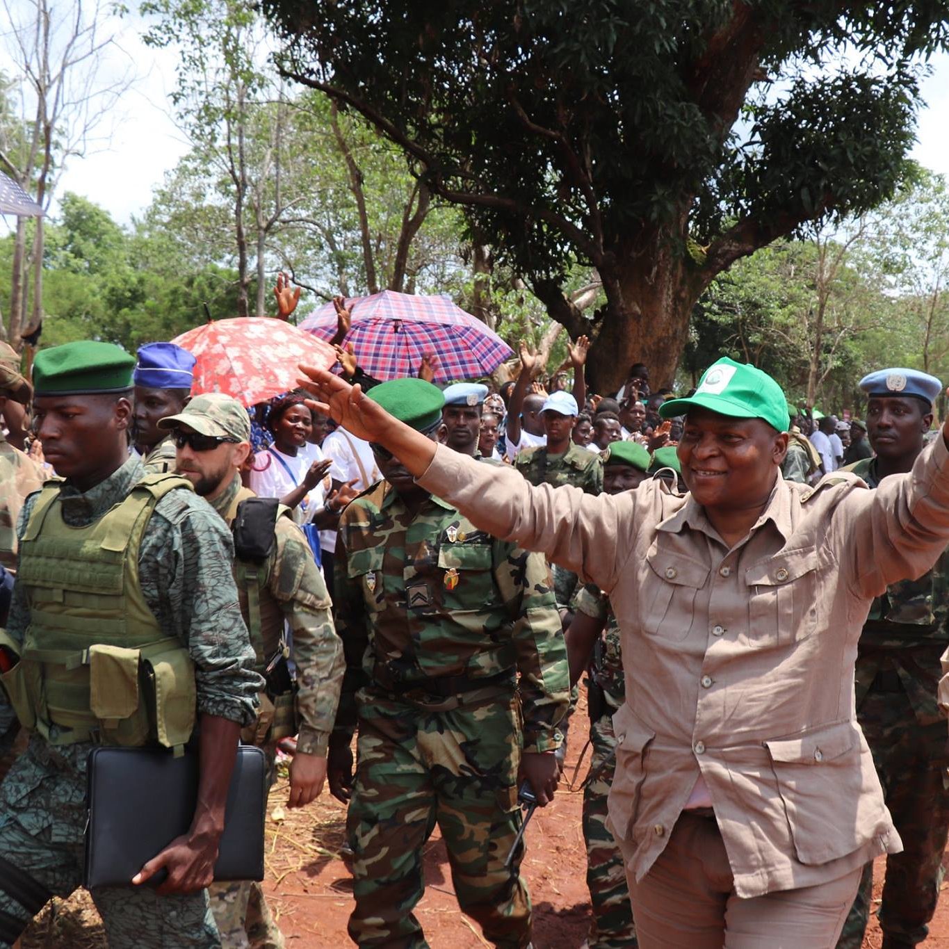 L'image montre une scène animée en plein air, avec un groupe de personnes réunies. Au centre, un homme souriant, vêtu d'une tenue beige et portant une casquette verte, semble saluer la foule avec les bras levés. Autour de lui, plusieurs militaires en uniforme, certains portant des gilets pare-balles, se tiennent en rang. La foule en arrière-plan est composée de personnes sous des parapluies, suggérant une atmosphère festive ou de célébration. Des arbres verdoyants et un chemin de terre sont visibles, créant un cadre naturel et vivant. L'ambiance semble énergique et encourageante.