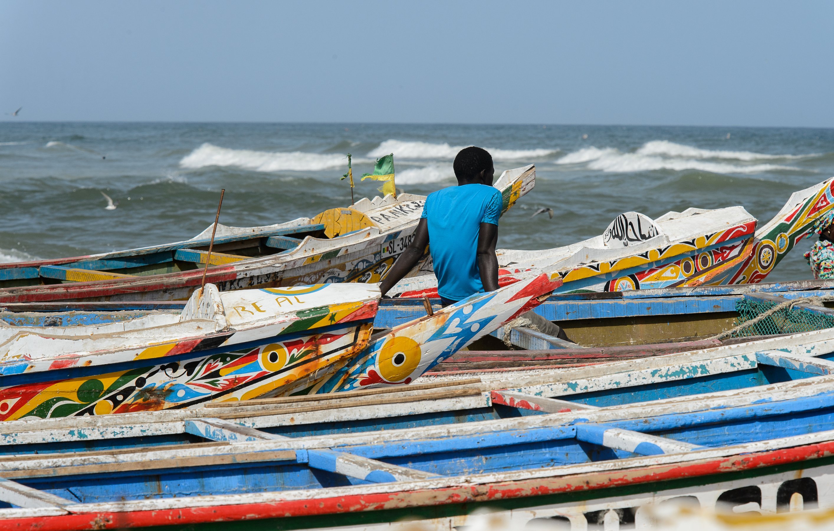 L'image montre une plage animée au bord de la mer. Des bateaux colorés se chevauchent sur le sable, avec des motifs vifs et des teintes variées, tels que le bleu, le rouge et le jaune. Un homme, vêtu d'un t-shirt bleu, est assis au bord des bateaux, tourné vers l'océan. Les vagues s'écrasent doucement sur le rivage, créant un bruit apaisant. Dans l'air, on peut sentir une brise marine fraîche et éventuellement entendre le cri des oiseaux qui volent autour. L'ambiance dégage une sensation de tranquillité tout en étant dynamique grâce à la présence des bateaux et de l'homme observant l'horizon.