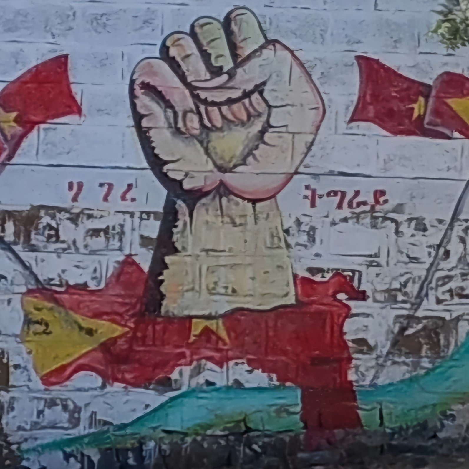 Sur le mur, on peut voir une grande fresque représentant un poing levé, symbole de résistance et de solidarité. Ce poing est peint dans des tons clairs, avec des détails qui soulignent sa force. Autour de lui, plusieurs drapeaux flottent, chacun arborant des motifs distincts en rouge et jaune, rappelant des symboles nationaux. En arrière-plan, on distingue des formes qui évoquent des paysages, créant une ambiance dynamique et engageante. L'ensemble transmet une forte impression de détermination et d'unité.