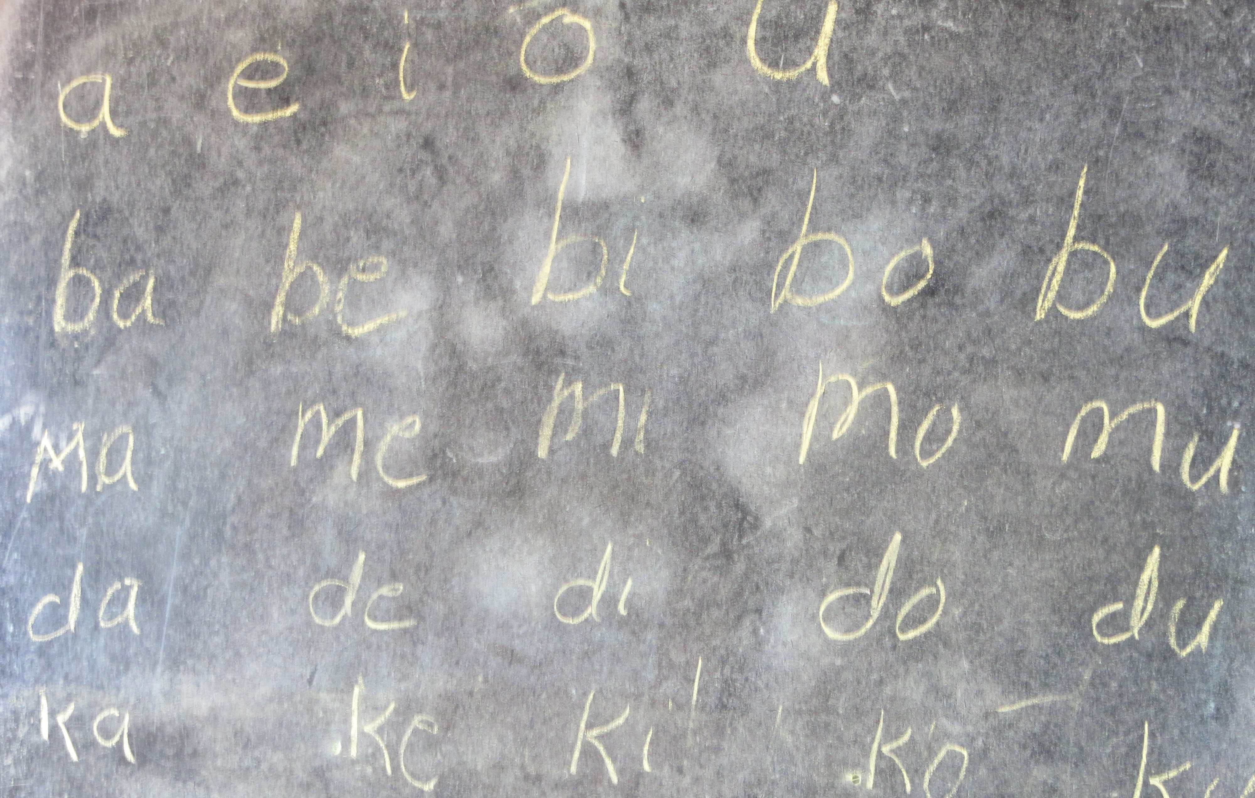 L'image montre un tableau noir sur lequel sont écrits des caractères en lettres cursives, à la craie. Les lignes commencent par les voyelles "a", "e", "i", "o", "u", suivies de combinaisons de consonnes et voyelles, telles que "ba", "be", "bi", "bo", "bu", puis "ma", "me", "mi", "mo", "mu", et ainsi de suite. L'écriture est claire et simple, et le tableau a une texture légèrement rugueuse. L'ensemble de l'image évoque un environnement d'apprentissage.