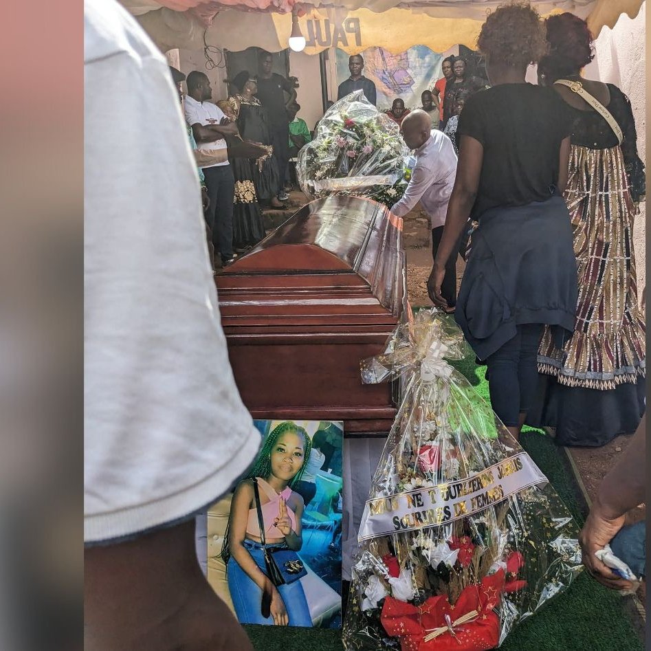 L'image montre un moment poignant d'un enterrement. Au centre, un cercueil en bois brun, sur lequel sont déposés des bouquets de fleurs. À côté du cercueil, une grande photo d'une femme est accrochée, entourée de décorations florales et de bandes de tissu. Des personnes, visiblement en deuil, se rassemblent autour, certaines portant des vêtements sombres. L'ambiance est sombre mais respectueuse, marquée par la tristesse et l'hommage à la personne décédée. L'arrière-plan est flou, mais on peut deviner une tente qui abrite cette cérémonie. L'ensemble évoque des sentiments de perte et de commémoration.