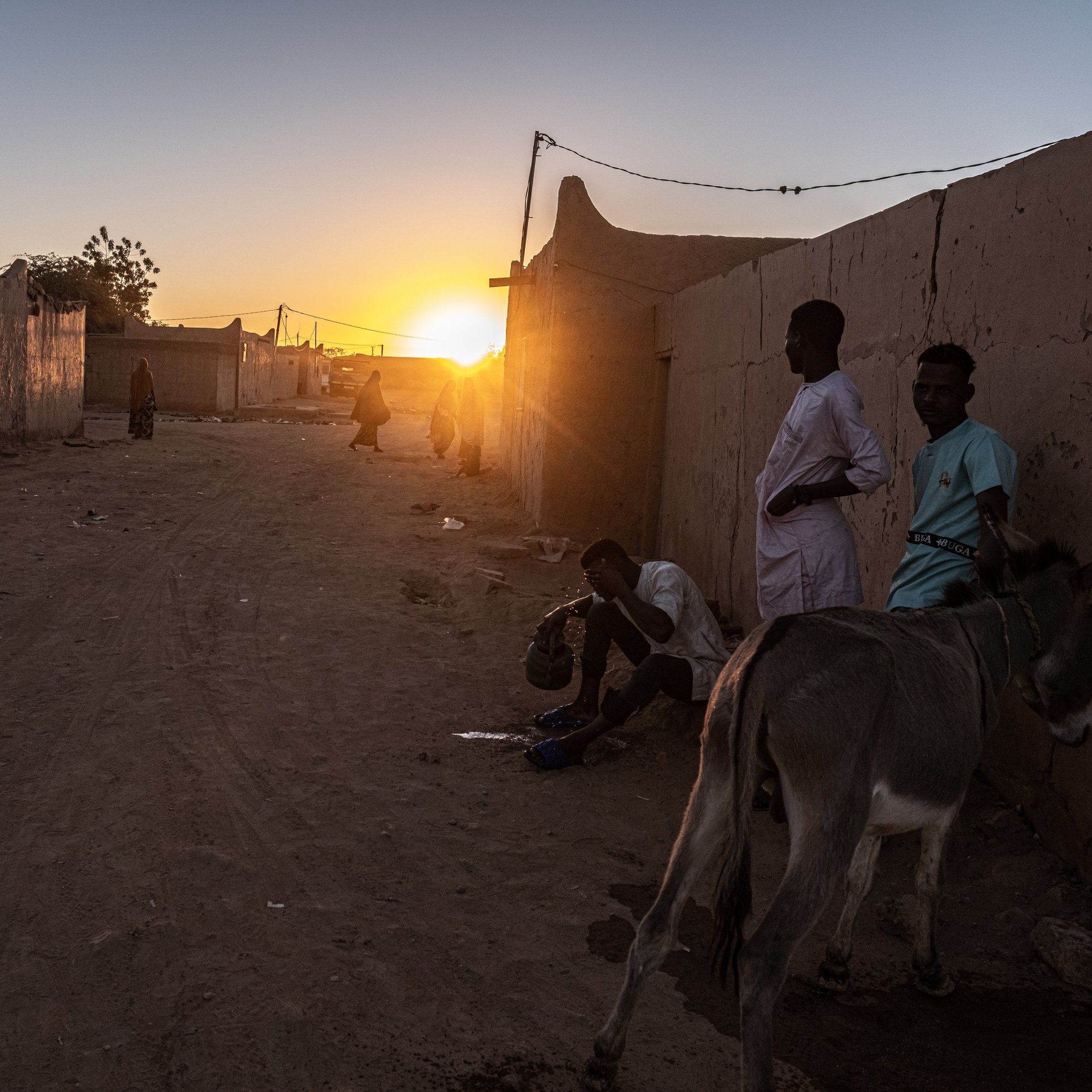Dans cette image, nous sommes confrontés à un paysage désertique au crépuscule. La lumière du soleil, qui descend à l'horizon, diffuse des teintes chaleureuses d'or et d'orange, illuminant doucement les bâtiments en terre ocre qui bordent la rue. À gauche, un âne se tient paisiblement, ajoutant une touche de vie à la scène. Sur le sol, la poussière et les cailloux témoignent d'une vie quotidienne simple. Des silhouettes d'hommes en vêtements traditionnels se déplacent lentement, discutant ou vaquant à leurs occupations, créant une atmosphère de convivialité et de calme. Les murs des maisons en terre, bien que rudimentaires, dégagent un sentiment de communauté. Dans l'ensemble, cette scène évoque une fin de journée tranquille, où les sons de la nature et les murmures des conversations se mêlent à la douce lumière du crépuscule.
