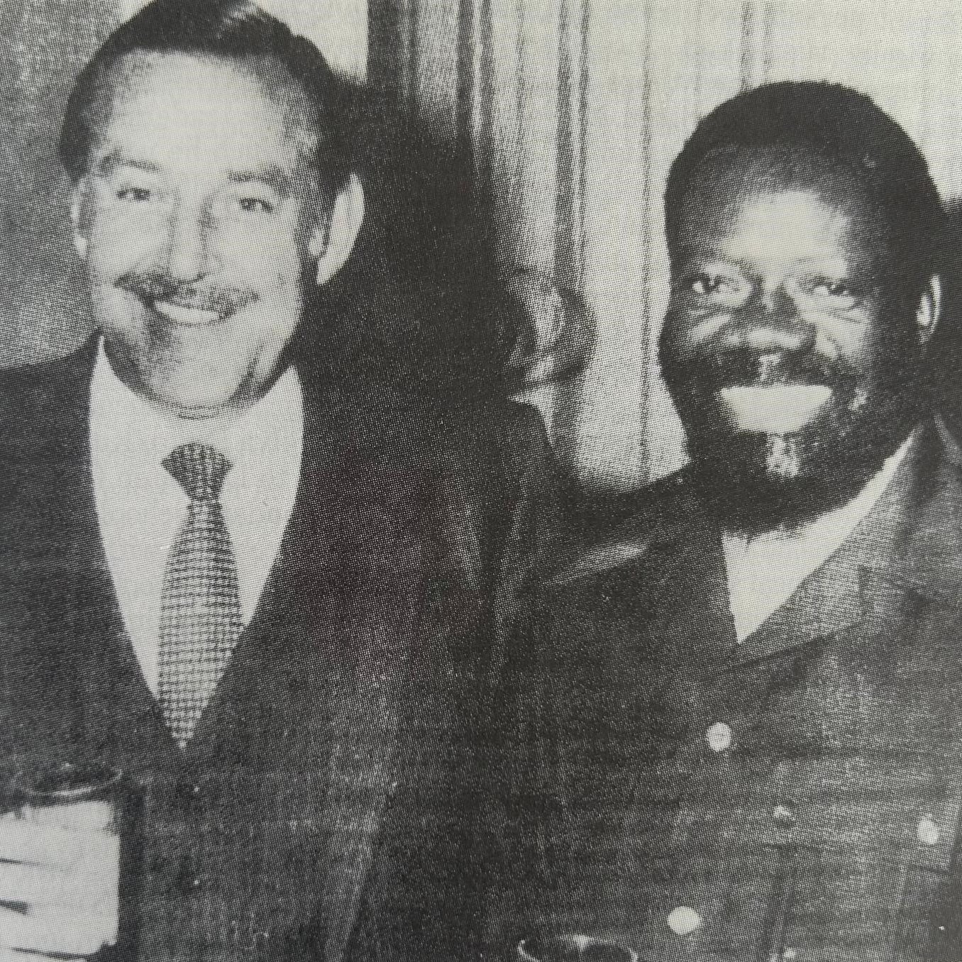 L'image montre deux hommes se tenant côte à côte, souriant largement. Le premier homme, à gauche, porte un costume sombre avec une cravate. Il a des cheveux grisonnants, une moustache bien dessinée et une expression amicale. Le deuxième homme, à droite, est habillé d'une veste de style militaire et a une barbe bien fournie. Il a les cheveux frisés et sourit également, ce qui donne une atmosphère chaleureuse à la photo. Les deux tiennent des verres à la main, suggérant une occasion conviviale, peut-être une célébration ou un événement. L'arrière-plan est flou, mettant l'accent sur les deux hommes.