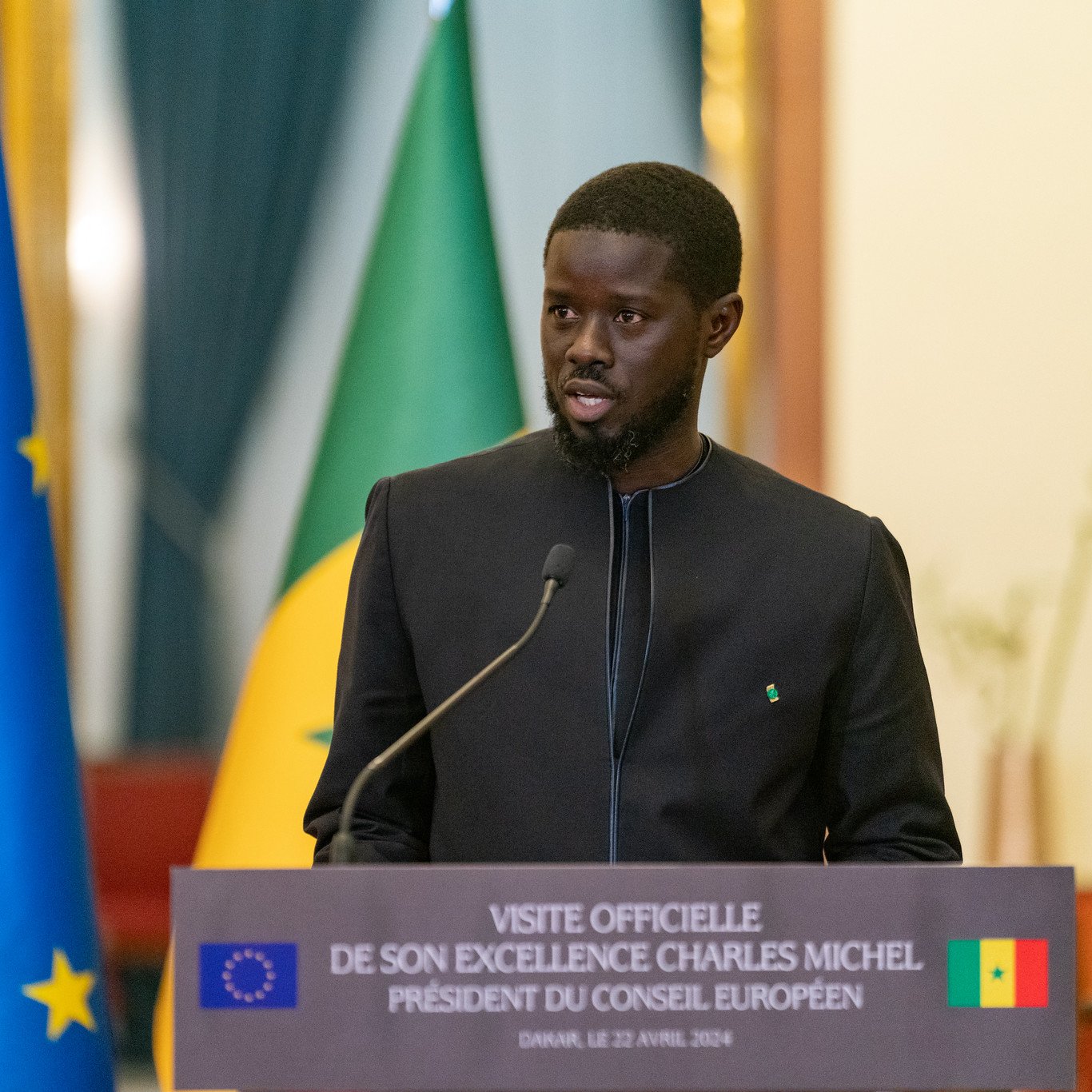 Sur cette image, un homme est en train de prendre la parole lors d'une conférence. Il est vêtu d'une tenue traditionnelle noire et se tient devant un podium portant une plaque avec des inscriptions officielles. En arrière-plan, on peut voir deux drapeaux : l'un affiche les étoiles de l'Union européenne et l'autre représente le Sénégal. L'homme semble sérieux et engagé, s'adressant à un public. La scène dégage une atmosphère formelle et diplomatique.