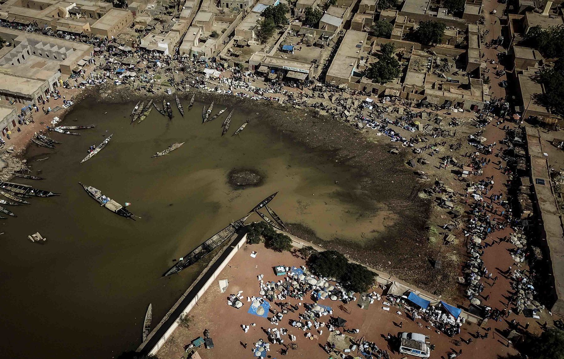 Imaginez un paysage vu du ciel. Au centre, un plan d'eau sombre qui semble être une lagune ou un port. Les bords de l'eau sont bordés de bateaux, certains amarrés, d'autres flottant paisiblement. Autour de cette lagune, il y a des habitations, des maisons en terre ou en tuiles, formant un petit village. Sur les rives, des gens sont rassemblés, certains assis par terre, d'autres se déplaçant avec des marchandises. Des tentes et des parasols sont disposés, suggérant un marché vivant où l'on troque et vend des produits. L'air est probablement rempli de sons, de cris et d'odeurs de nourriture, de poissons et de marchandises variées, donnant l'impression d'une scène animée et prospère. Les couleurs sont chaudes, évoquant un climat ensoleillé. Ce tableau dégage une atmosphère d'activité et de communauté autour de l'eau.