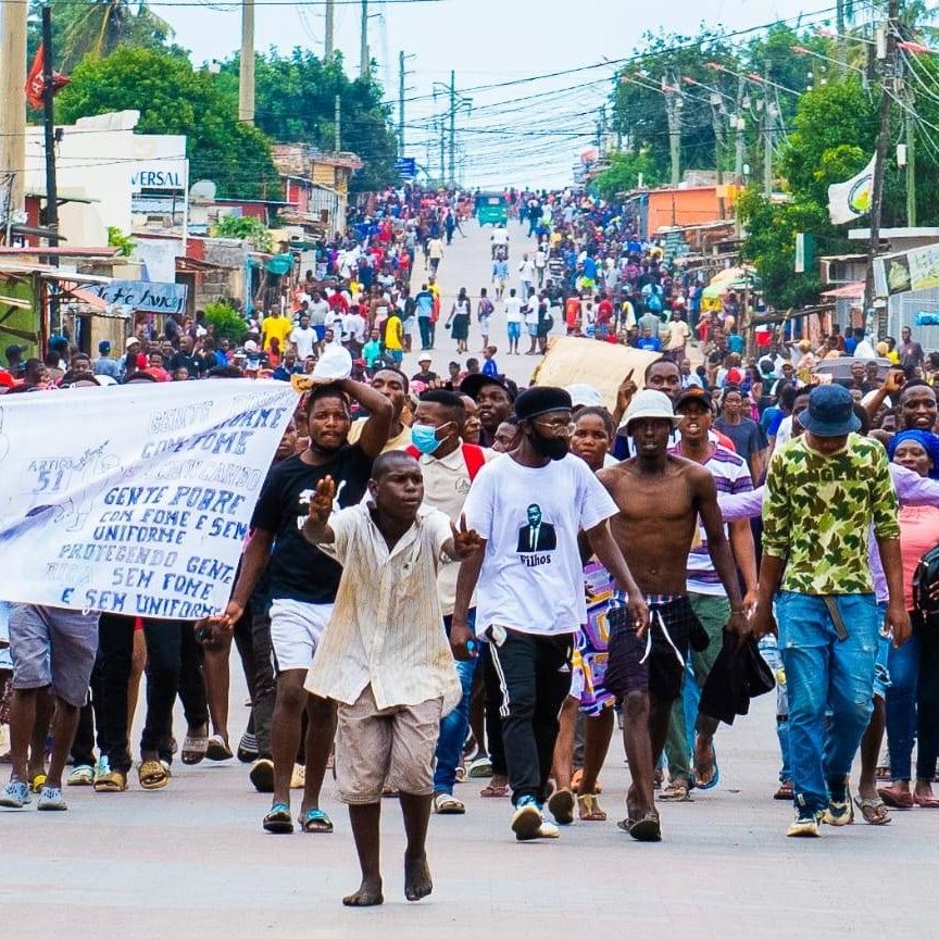 L'image montre une foule nombreuse marchant dans une rue animée. Les gens sont regroupés, certains tiennent des banderoles ou des pancartes. L'ambiance semble dynamique, avec beaucoup de mouvements et des échanges d'expressions entre les participants. On peut également ressentir une diversité dans les vêtements, allant des tenues décontractées aux vêtements plus colorés. En arrière-plan, des boutiques et des bâtiments bordent la rue, tandis que des fils électriques sont suspendus au-dessus. Les voix et les chants des manifestants semblent animer l'espace, créant une atmosphère de rassemblement et de mobilisation.