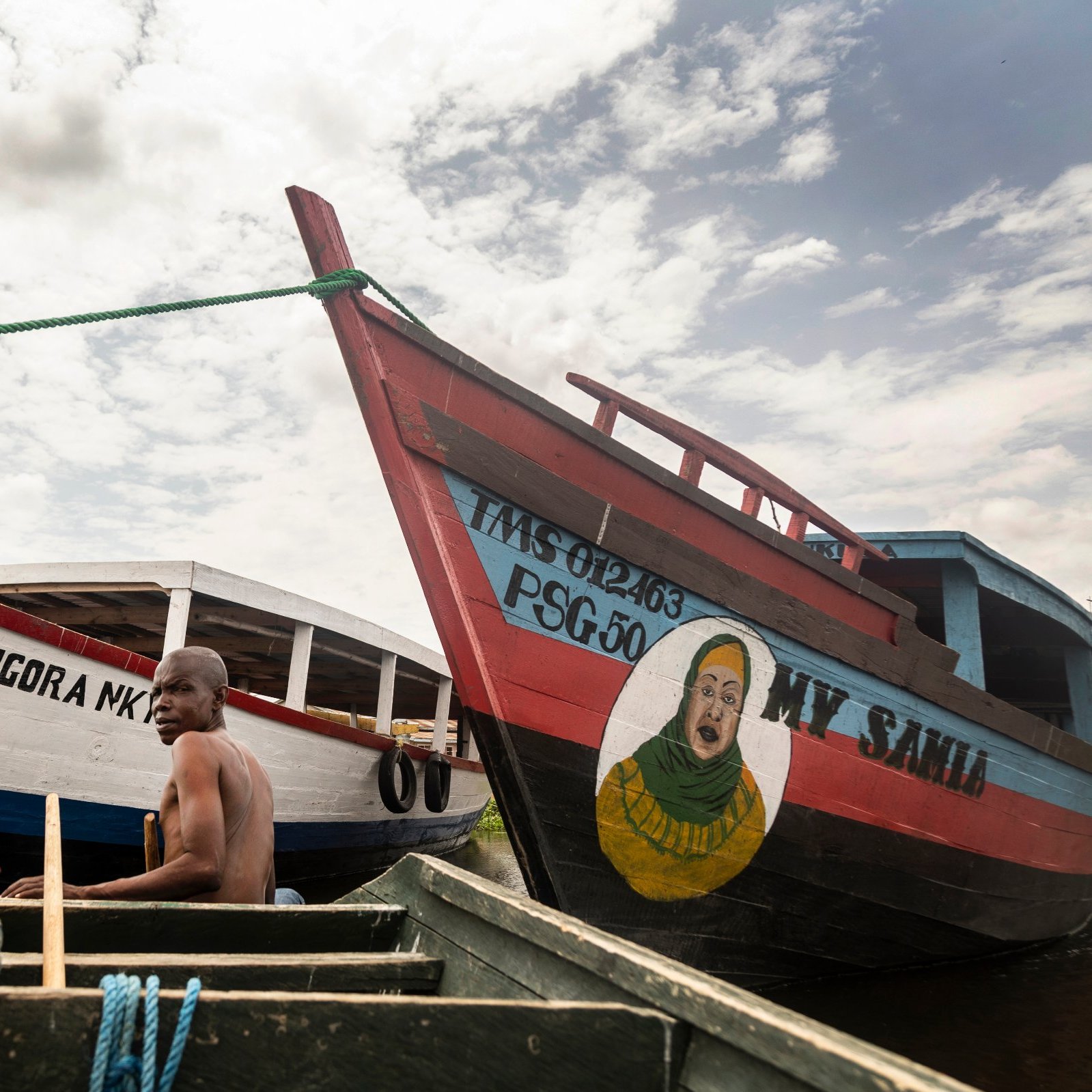 L'image montre deux bateaux colorés amarrés au bord d'un cours d'eau calme. À l'avant, un homme, en train de ramer, est visible, concentré sur son effort. Un autre homme est assis à l'arrière, regardant vers l'objectif. Les bateaux sont peints de manière vive, avec des inscriptions comme "MY SAID" et "MONGORA MK". L'un des bateaux présente également une grande peinture d'un visage souriant. Le ciel est partiellement nuageux, ajoutant une dimension lumineuse à la scène. L'atmosphère est paisible, évoquant une ambiance de vie flottante.