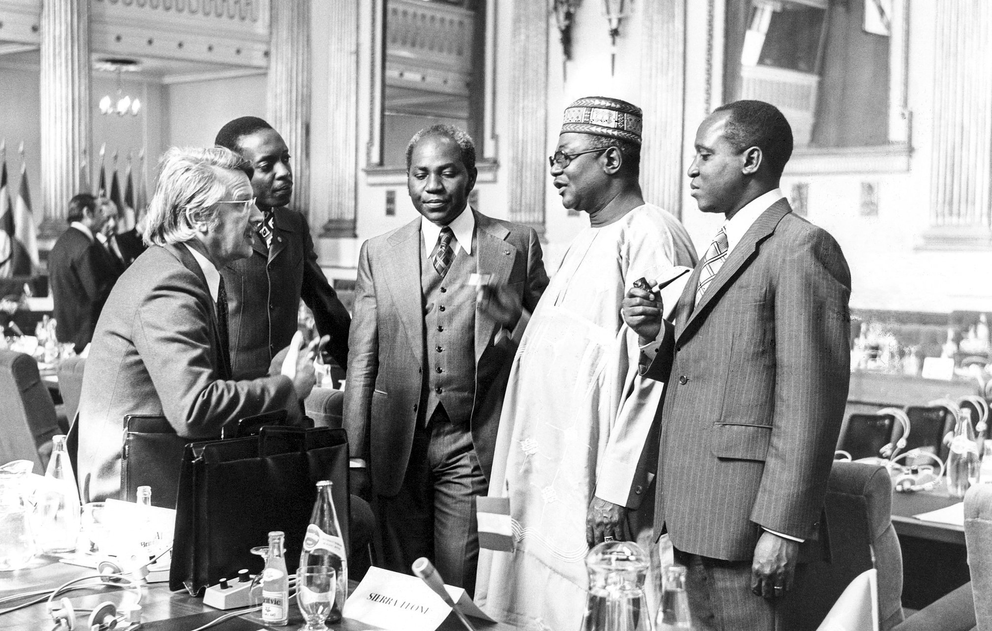 L'image représente un groupe de cinq hommes en discussion dans un cadre formel, probablement lors d'une conférence ou d'une réunion. Ils sont habillés en costumes élégants, dont certains sont accompagnés de cravates. Les visages montrent une interaction animée, avec des gestes de mains qui suggèrent une conversation engageante. En arrière-plan, on aperçoit des drapeaux, ce qui indique un contexte international. La scène se déroule dans une grande salle, ornée et bien éclairée, ajoutant une atmosphère de sérieux et de respect.