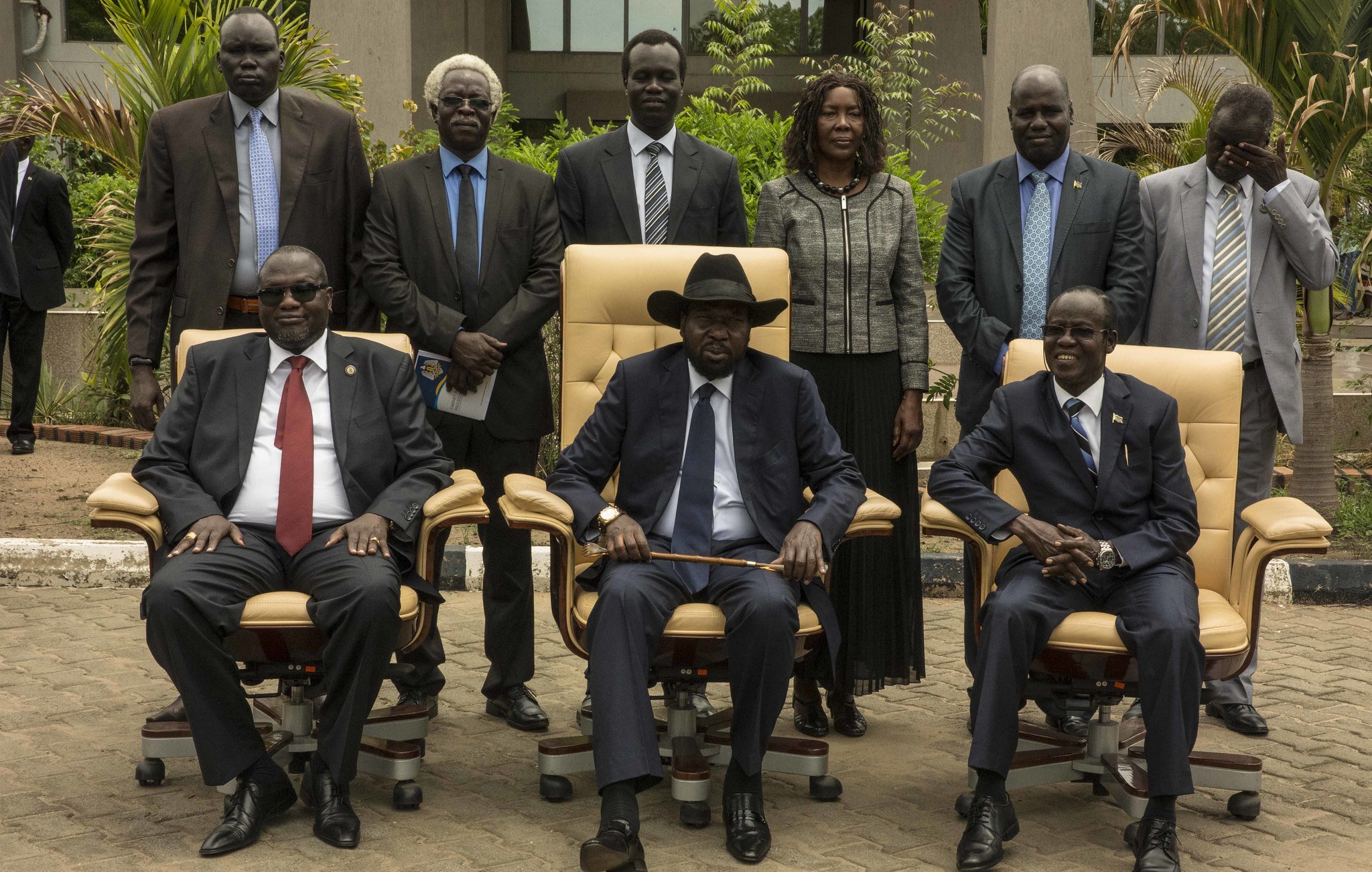 L'image présente un groupe de personnes posant ensemble à l'extérieur d'un bâtiment. Au centre, on voit trois hommes assis dans de grandes chaises confortables. Le premier homme à gauche porte des lunettes, un costume sombre et une cravate rouge. Le second, au centre, est vêtu d'un costume noir, d'une cravate et d'un chapeau noir, et semble être le leader du groupe. Le troisième homme, à droite, porte également un costume et une cravate. Derrière eux, plusieurs personnes se tiennent debout, toutes en tenue formelle. L'environnement est verdoyant, avec des plantes et un chemin en pavé. L'atmosphère semble sérieuse et officielle, suggérant une rencontre importante.