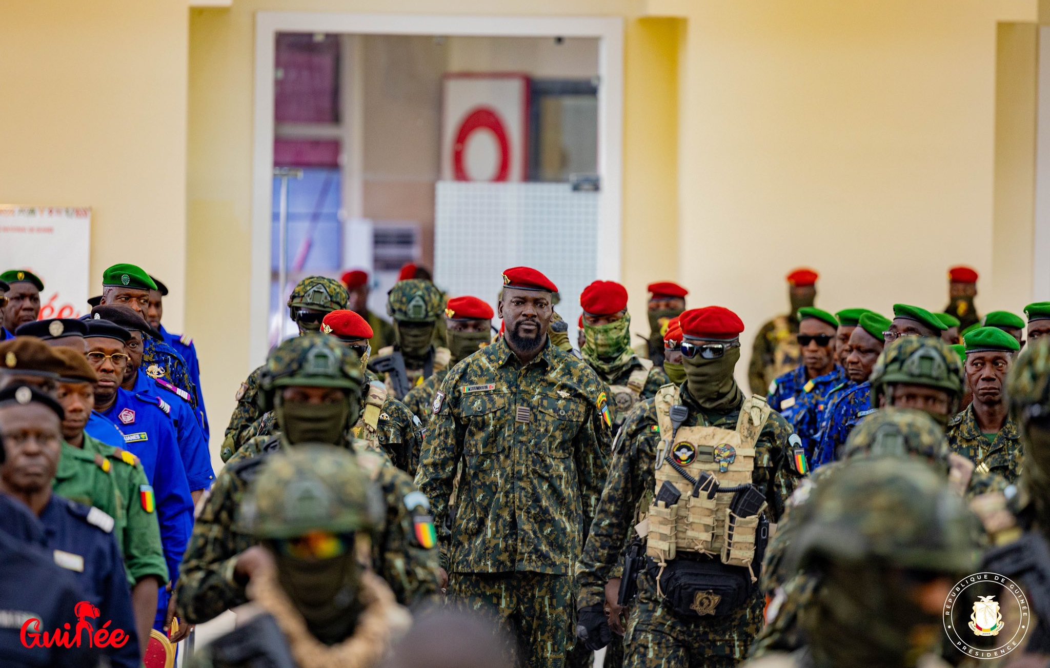L'image représente un groupe de militaires rassemblés dans un intérieur lumineux. Au centre, un homme en uniforme se distingue par son attitude imposante. Il porte un camouflage militaire et un béret rouge. Les autres soldats autour de lui sont vêtus d'uniformes similaires, certains portant des masques de camouflage. Leurs bérets varient en couleur, notamment le rouge et le vert, créant un contraste visuel. L'ensemble évoque une atmosphère de discipline et de solidarité au sein des forces armées.