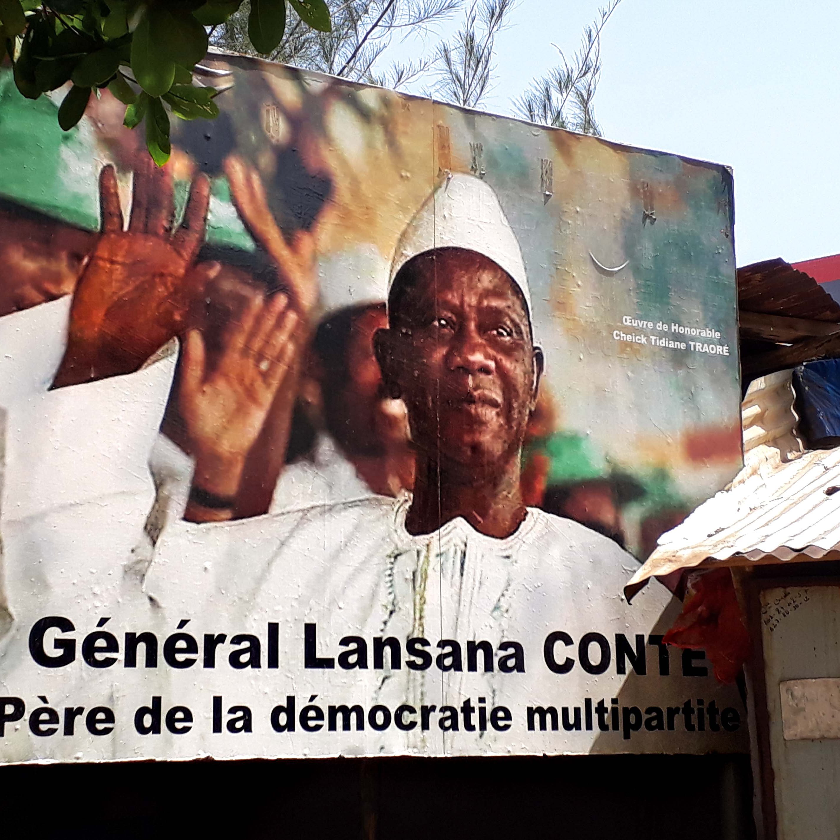 L'image représente une grande affiche murale d'un homme en train de saluer, avec un sourire bienveillant. Il porte une tenue traditionnelle, incluant un chapeau blanc et une chemise claire. En arrière-plan, on peut voir des couleurs vives qui semblent évoquer une atmosphère festive ou patriotique. Au bas de l'affiche, le texte indique « Général Lansana Conté, Père de la démocratie multipartite », soulignant ainsi son rôle important dans l'histoire politique. Les lignes et les formes des éléments de l'affiche créent un contraste qui attire le regard, suggérant un message de respect et d'honneur envers cette figure politique.