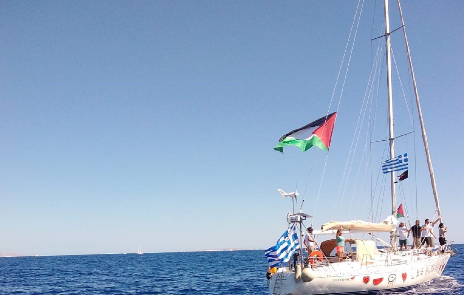 L'image présente un voilier sur une mer turquoise calme, sous un ciel d'un bleu éclatant sans nuages. Le bateau, de couleur blanche, arbore plusieurs drapeaux, dont celui de la Palestine, de la Grèce, et d'autres drapeaux en bandes horizontales typiques. À bord, on aperçoit des personnes qui semblent profiter d'une journée en mer. Les vagues légères créent une ambiance paisible, et l'horizon lointain s'étend, avec d'autres bateaux visibles au loin. L'ensemble évoque une atmosphère de liberté et d'aventure maritime.