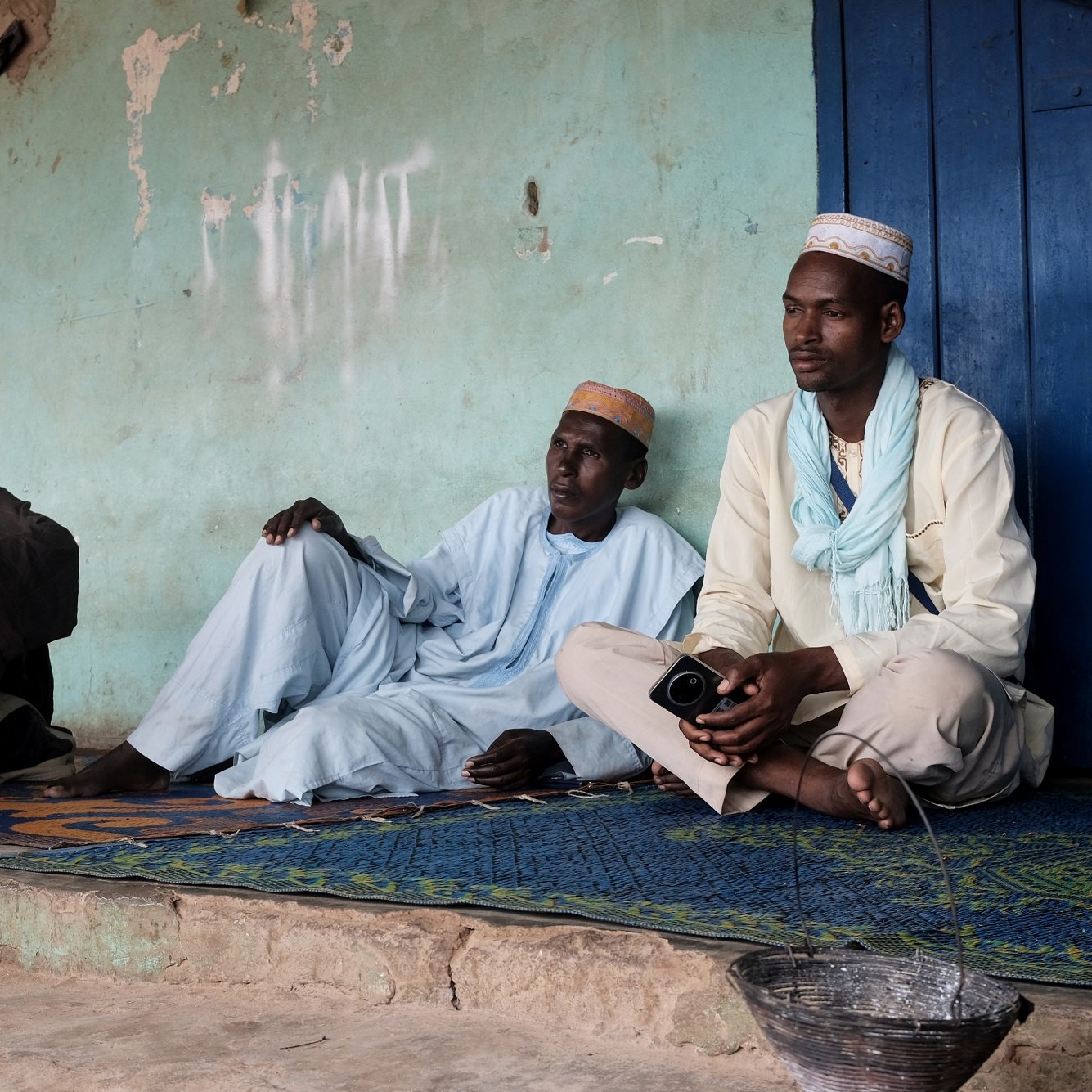 Cette image montre trois hommes assis sur un tapis, à l'extérieur d'un bâtiment aux murs de couleur turquoise. L'atmosphère semble calme et détendue. Ils portent des vêtements traditionnels : deux d'entre eux sont vêtus de tuniques, tandis que le troisième porte un chapeau. L'un des hommes est assis en tailleur, tandis que les deux autres sont en position plus détendue, le dos appuyé contre le mur. À côté d'eux, il y a un petit seau en métal d'où s'écoule un peu d'eau. En arrière-plan, des murs sont partiellement recouverts de graffiti. L'environnement semble rural, avec une lumière douce qui illumine la scène.