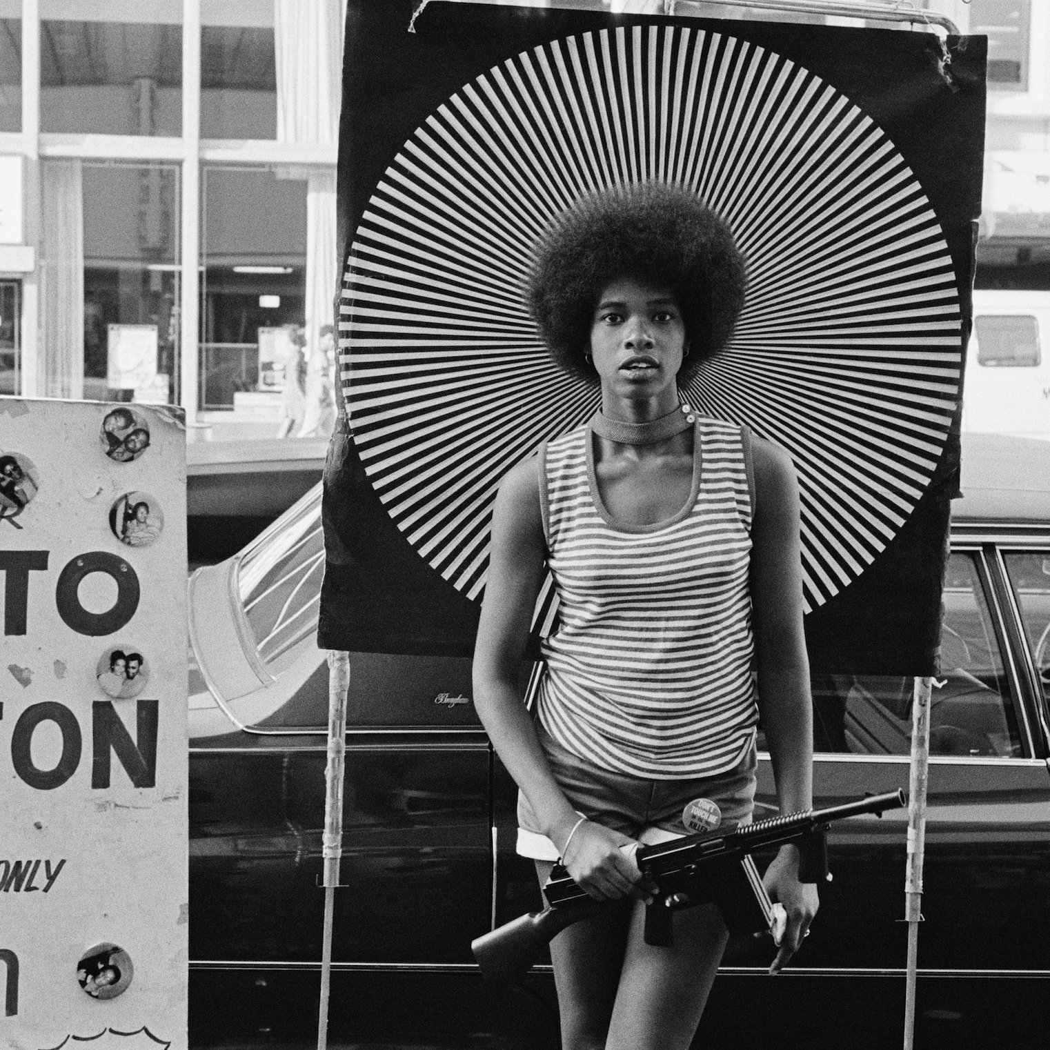 L'image présente une scène urbaine en noir et blanc. Au premier plan, une jeune femme avec une coiffure afro se tient de manière confiante, tenant une arme à feu. Elle porte un haut à rayures et se tient devant un arrière-plan décoré de motifs en spirale noirs et blancs, ce qui attire l'œil et crée un contraste frappant. À sa gauche, un panneau indique "PUT YOUR PHOTO ON A BUTTON" avec du texte dans une police simple, ajoutant un contexte de marché ou d'événement communautaire. En arrière-plan, on peut apercevoir des bâtiments et des véhicules, suggérant une atmosphère animée de la ville. L'image évoque des thèmes de pouvoir, de culture et de contestation.