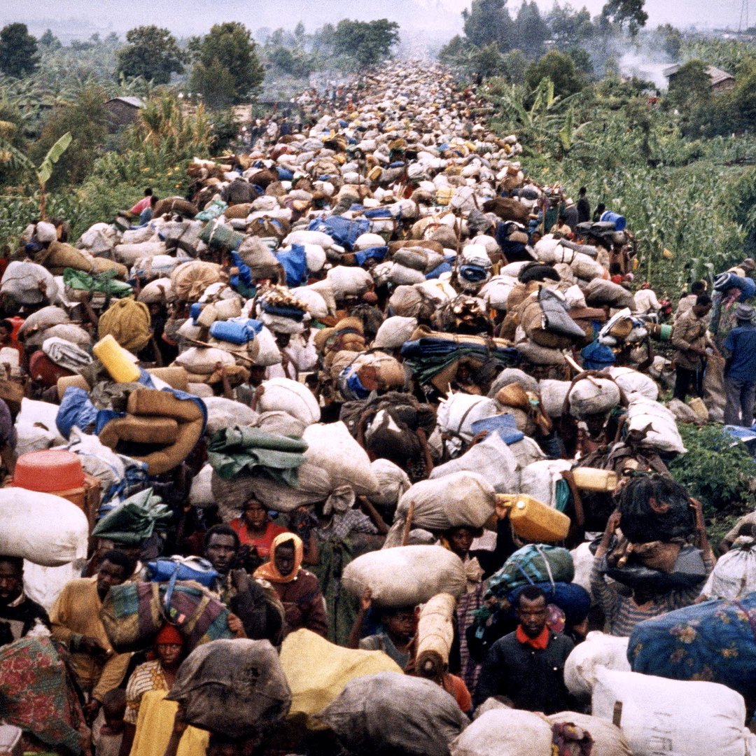 L'image montre une scène de déplacement massifs de personnes. Une foule dense de réfugiés marche sur un chemin, transportant de lourdes charges sur le dos et la tête. Les personnes sont entourées de verdure, avec des arbres et des plantations visibles au loin. Les visages des réfugiés reflètent souvent la fatigue et la détermination, tandis que certains portent des vêtements colorés. L'ambiance est marquée par un sentiment de lutte et de solidarité, malgré les conditions difficiles de leur voyage. Le paysage est ombragé par des montagnes en arrière-plan, ajoutant à la profondeur de la scène.