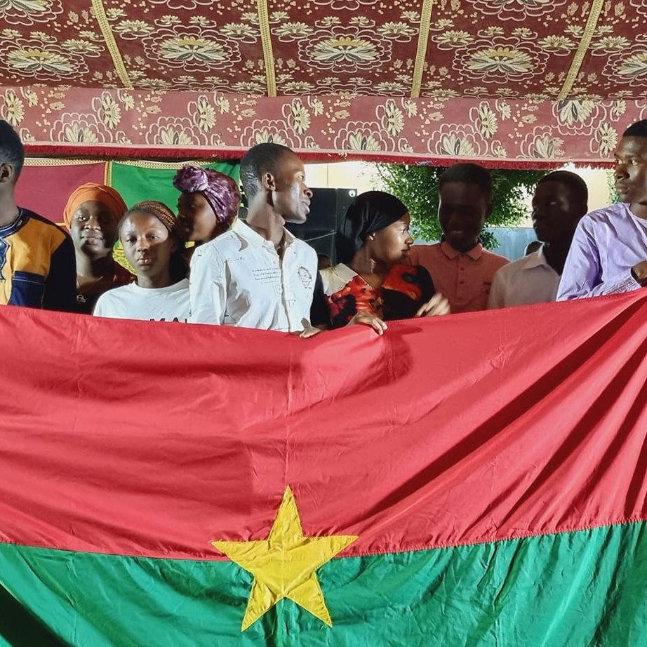 L'image présente un groupe de jeunes personnes rassemblées sous une tente décorée, avec des motifs colorés en arrière-plan. Au centre, ils tiennent une grande drapeau du Burkina Faso, qui est rouge avec une étoile verte au milieu. Les membres du groupe affichent des vêtements variés, certains portant des chemises traditionnelles, et semblent joyeux et solidaires. L'atmosphère est festive, suggérant un événement culturel ou une célébration.