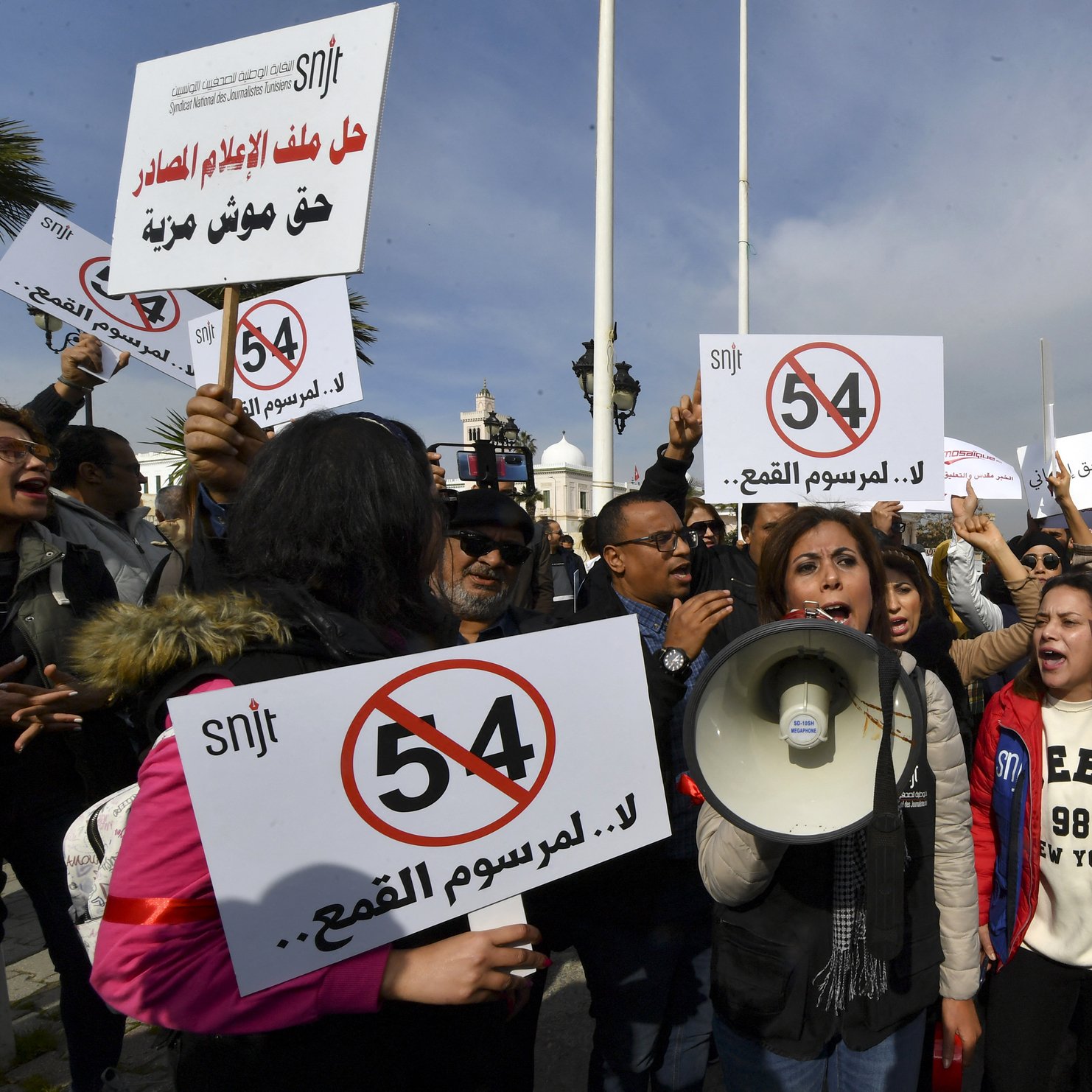 The image depicts a protest scene with a group of people holding signs and banners. In the foreground, a woman is speaking into a megaphone, likely leading the demonstration. The signs feature the number "54" and messages opposing censorship, suggesting that the protest is focused on issues related to freedom of expression or press freedom. The crowd appears to be actively engaged in the protest, raising their signs and voicing their demands. The setting seems to be outdoors, with flags and possibly a government building in the background.
