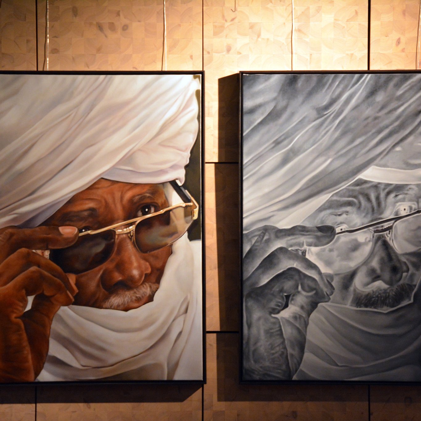 L'image présente deux tableaux accrochés sur un mur, représentant le même homme, mais dans des styles différents. À gauche, le portrait est en couleurs vives : un vieil homme porte un turban blanc et des lunettes de soleil. Son regard est pensif, avec une expression de sagesse. À droite, l'œuvre est en noir et blanc, offrant un contraste fort. Les détails des rides et des textures de la peau sont accentués, donnant une sensation de profondeur. En bas à droite de l'image, on aperçoit une silhouette d'un visiteur qui observe les œuvres. Le fond du mur est constitué de panneaux en bois clair, ajoutant une dimension chaleureuse à l'espace d'exposition.