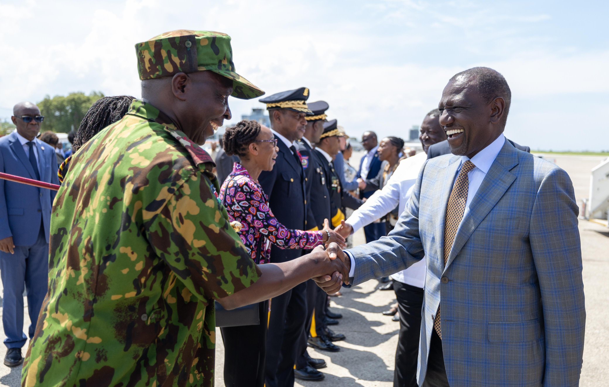 L'image montre une scène de bienvenue officielle à l'aéroport. Au premier plan, deux hommes se serrent la main, exprimant une atmosphère de camaraderie et de respect. L'un des hommes est en uniforme militaire avec un motif camouflage, tandis que l'autre porte un costume élégant avec une cravate. Autour d'eux, un groupe de personnalités, dont des membres des forces de sécurité et des fonctionnaires en civil, se tient en ligne, attendant leur tour pour saluer. En arrière-plan, le ciel est clair et ensoleillé, créant une ambiance positive. L'ensemble de la scène respire la solennité et l'importance d'un événement officiel.
