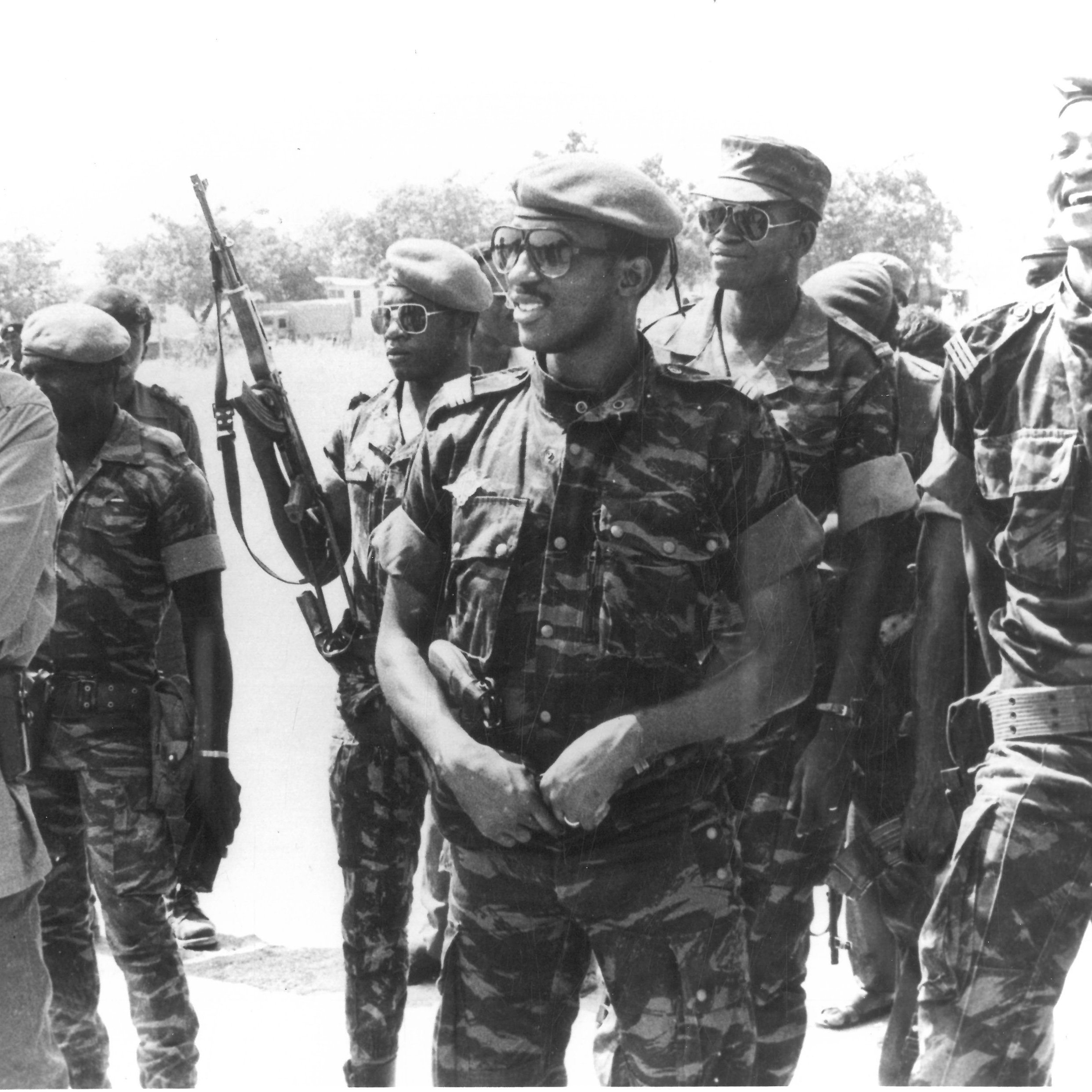 Cette image en noir et blanc montre un groupe de soldats en tenue militaire. Au premier plan, un homme souriant, portant un béret et une tenue de camouflage, se tient à gauche. À sa droite, plusieurs autres soldats, également en uniforme, affichent des expressions de camaraderie. L'un d'eux porte des lunettes de soleil et un autre semble être en train de rire. En arrière-plan, on peut voir des armes accrochées en position debout. L'ambiance générale de l'image suggère une atmosphère de camaraderie et de cohésion parmi les soldats.
