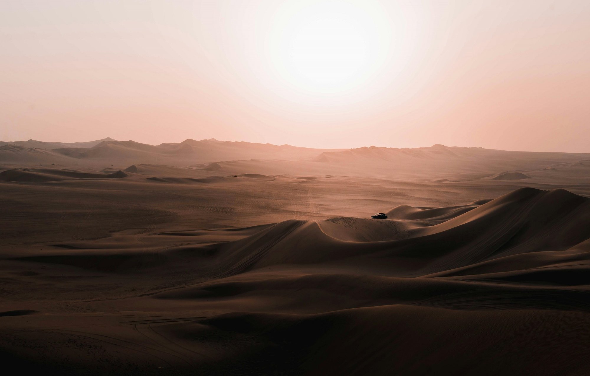 Imaginez une vaste étendue de sable, où les dunes s'étirent à perte de vue, leurs formes ondulantes créant des ombres douces sur le sol. Leurs contours se dessinent en vagues délicates, semblant danser sous les rayons d'un soleil flamboyant qui se lève à l'horizon, infusant le paysage d'une lumière chaude et dorée. Le silence règne, ponctué seulement par le léger murmure du vent qui balaie le désert, et une sensation de calme profonde imprègne l'air. Au loin, on devine une silhouette, peut-être un véhicule, qui semble minuscule face à l'immensité de ce panorama naturel. L'ensemble évoque une beauté brute, une tranquillité presque contemplative, comme si le temps s'était arrêté.