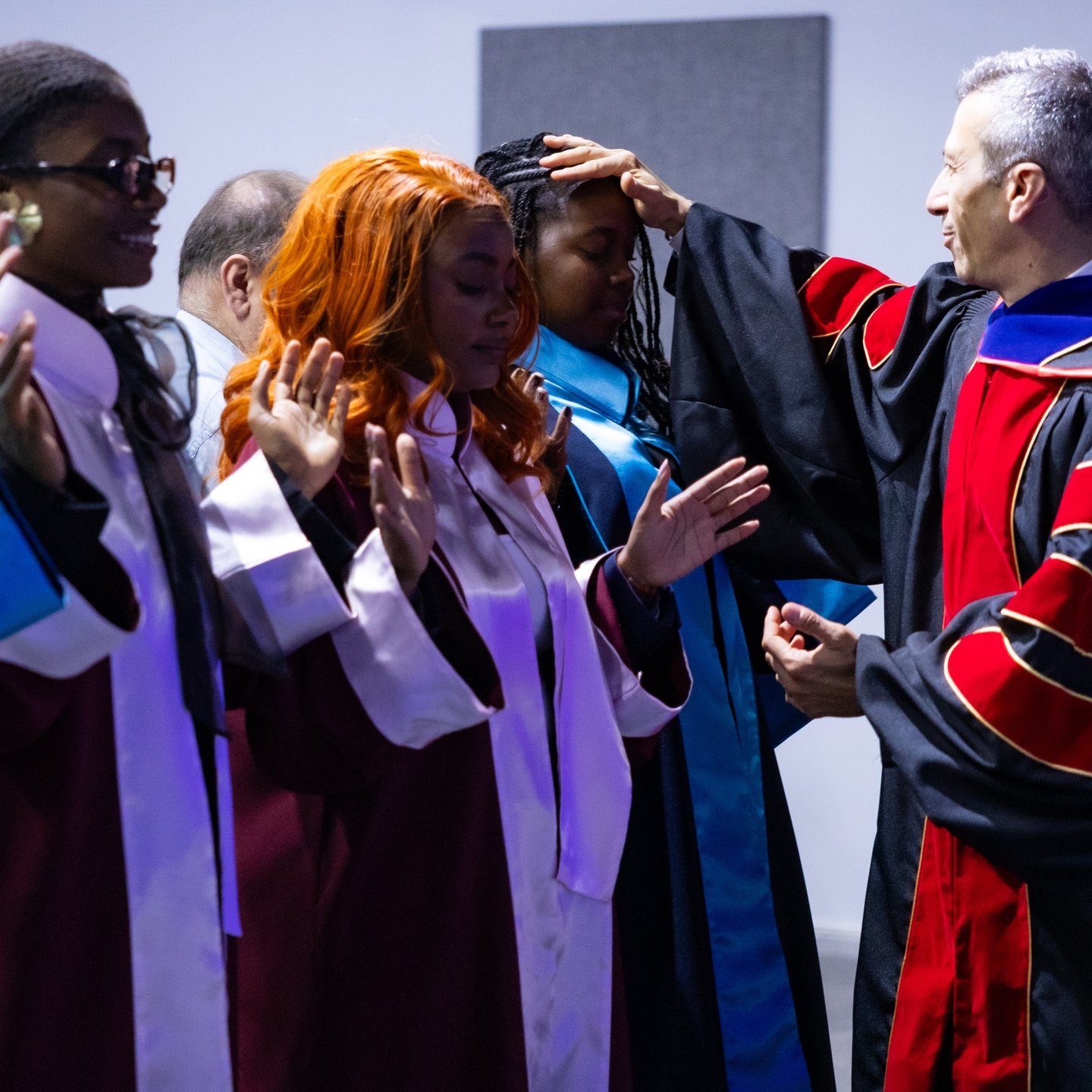 L'image montre une scène de cérémonie de remise des diplômes dans un auditorium. Plusieurs diplômés sont présents, portant des toges de différentes couleurs : certaines en bordeaux et d'autres en bleu. Ils sont debout en ligne, avec des expressions qui expriment à la fois la fierté et l'anticipation. Un homme, vraisemblablement un responsable de l'établissement, se tient devant eux, en train de poser une main sur la tête d'une diplômée avec des cheveux bouclés et roux. Les autres diplômés lèvent les mains, soit pour montrer leur gratitude, soit en signe de célébration. En arrière-plan, des personnes supplémentaires assistent à l'événement, complétant l'atmosphère festive de cette occasion mémorable.