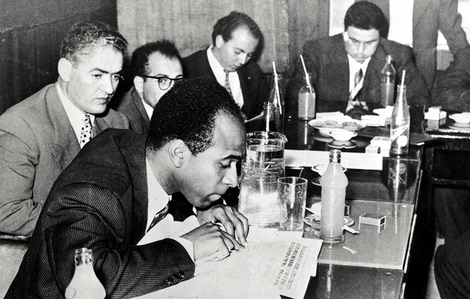 L'image en noir et blanc montre une scène de réunion dans un environnement intérieur, probablement une salle de conférence. Plusieurs hommes sont assis autour d'une table. Au premier plan, un homme est penché sur une feuille de papier, prenant semble-t-il des notes. Il est vêtu d'un costume sombre et d'une cravate, et semble concentré. À sa droite, un autre homme regarde vers le bas, tandis que les autres hommes en arrière-plan discutent ou examinent des documents. Sur la table, on peut voir des verres, des bouteilles, et quelques objets éparpillés. L'atmosphère semble sérieuse et studieuse, évoquant un moment important de collaboration ou de décision.