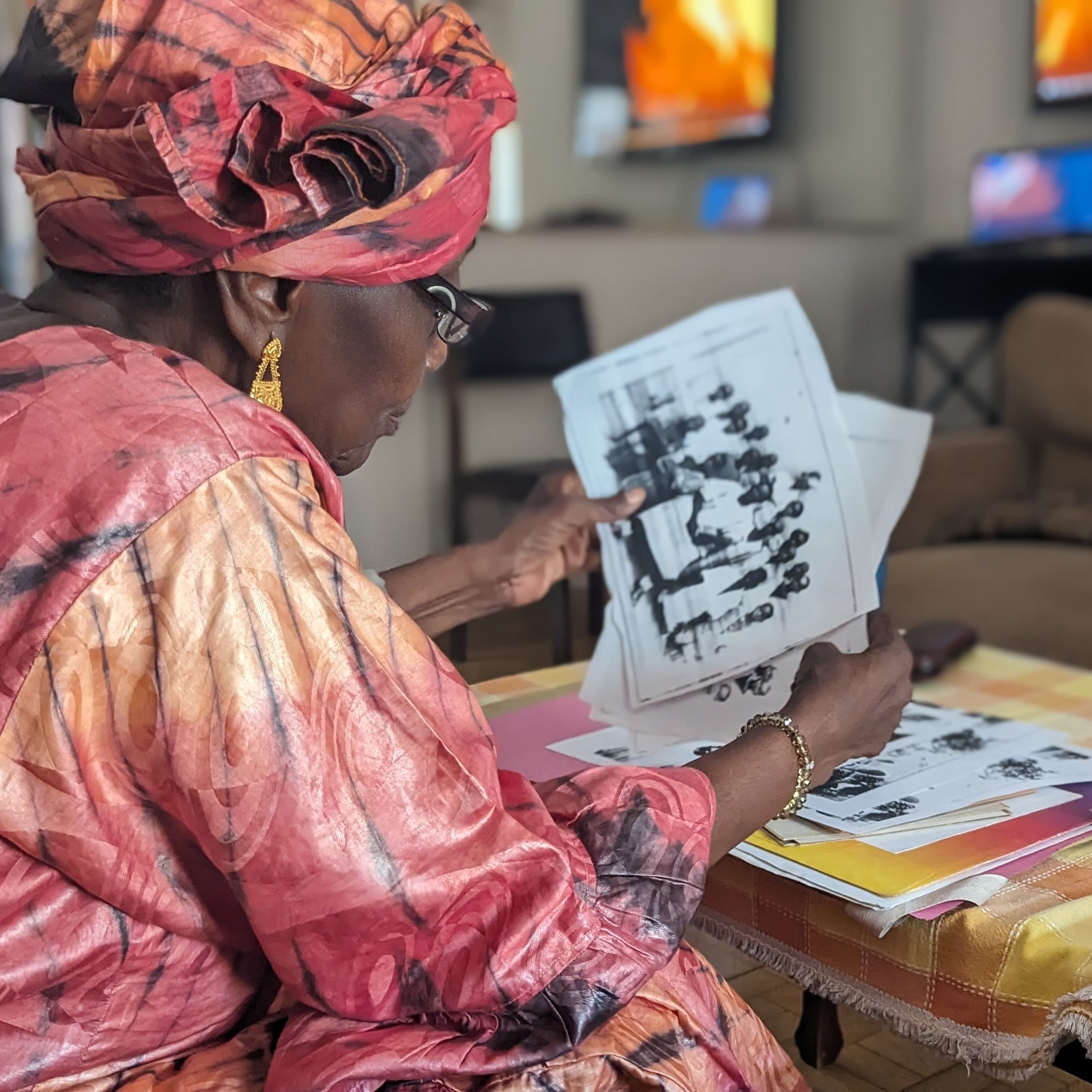 Dans cette image, une femme d'âge mûr, vêtue d'une magnifique robe rose et d'un foulard assorti, est assise sur un canapé dans un salon lumineux. Elle feuillette attentivement des photos en noir et blanc, toutes étalées devant elle sur une table basse recouverte d'une nappe à carreaux jaune et blanc. Le décor du salon est moderne, avec des télévisions visibles en arrière-plan, reflétant une ambiance chaleureuse et accueillante. La concentration de la femme témoigne de l'importance des souvenirs évoqués par ces images.
