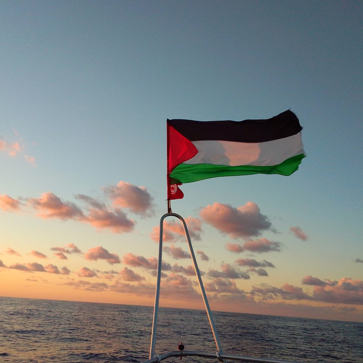 The image depicts a flag flying on a boat against a backdrop of a colorful sky during sunset. The flag features three horizontal stripes: black on the top, white in the middle, and green on the bottom, with a red triangle on the left side. The ocean can be seen below, and there are clouds in the sky that are illuminated by the setting sun, creating a serene atmosphere.