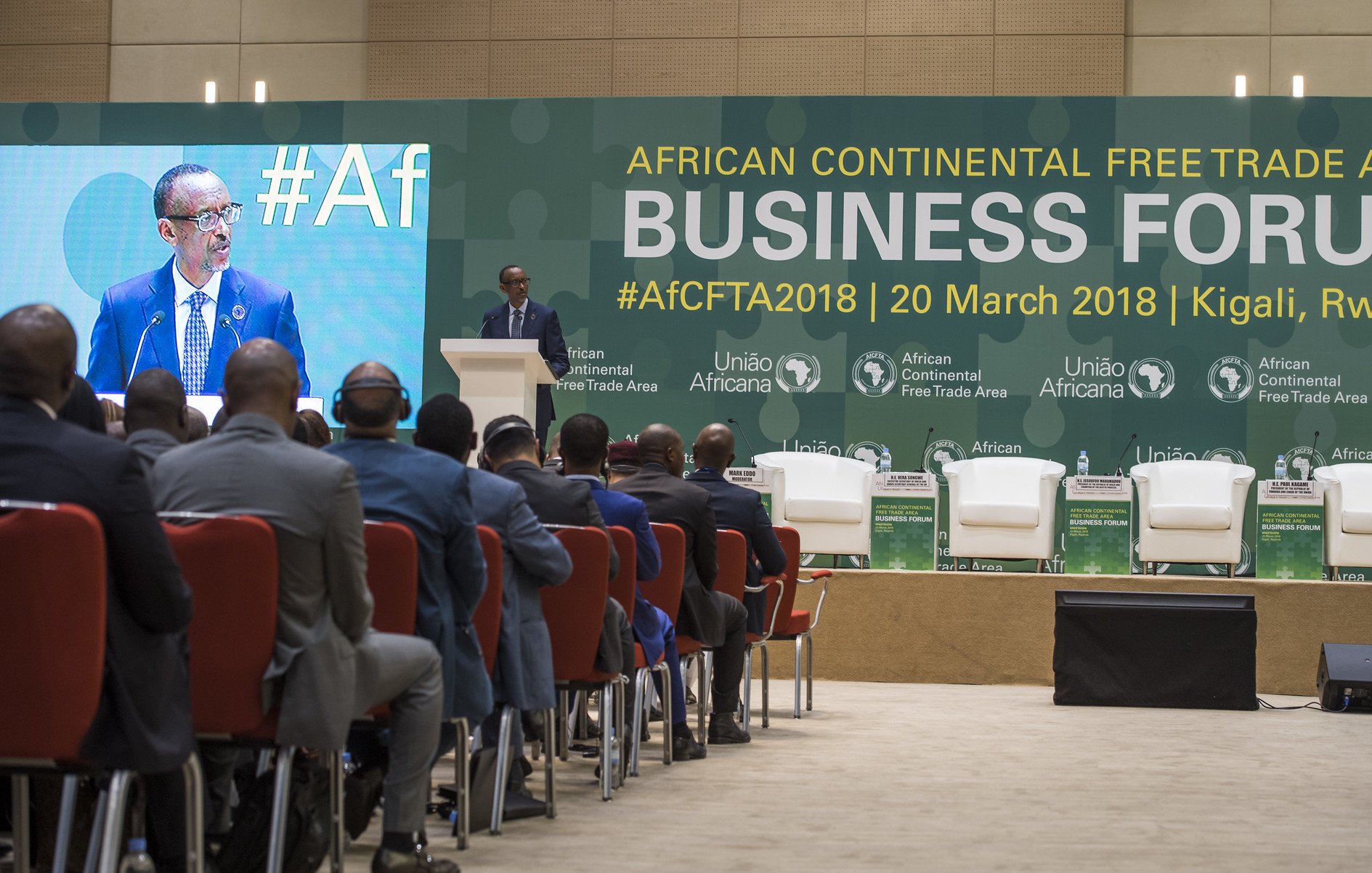 L'image présente un forum d'affaires qui se tient dans le cadre de la Zone de libre-échange continentale africaine. Au premier plan, on voit un homme, vraisemblablement un orateur, debout au micro, s'adressant au public. Il est entouré d'une scène bien agencée avec plusieurs chaises blanches et des logos des organisations impliquées dans l'événement. Le public, de dos, est composé d'hommes en costume, attentifs à la présentation. L'ambiance semble formelle et axée sur les discussions économiques. En arrière-plan, des banderoles mentionnent le forum et sa date, soulignant l'importance de l'événement pour le commerce en Afrique.