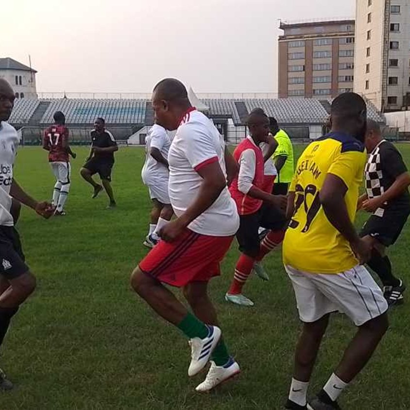 L'image représente un groupe de personnes jouant au football sur un terrain en herbe. Les joueurs portent des maillots de différentes couleurs, principalement blancs, rouges et jaunes. On peut voir des hommes en train de s'échauffer ou de danser, avec des mouvements dynamiques et joyeux. En arrière-plan, on aperçoit des gradins remplis, et des bâtiments se dressent autour du terrain. L'ambiance semble conviviale et énergique, avec un sentiment de camaraderie parmi les joueurs.