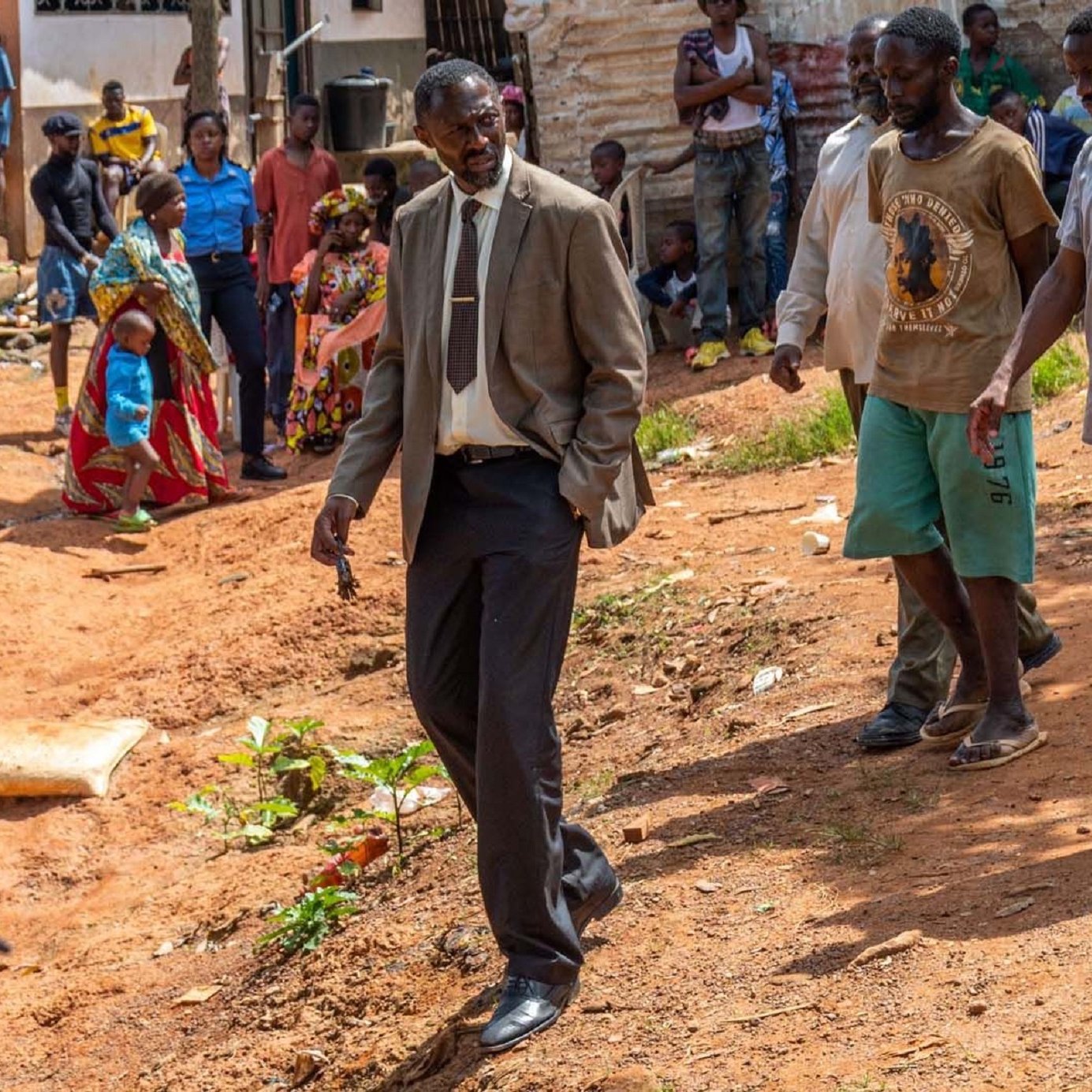 L'image montre un groupe de personnes dans un environnement rural ou semi-urbain. Au premier plan, un homme en costume cravate semble marcher avec assurance, le regard concentré. À sa gauche, un homme portant un t-shirt et un short se tient à côté de lui. D'autres personnes, habillées de manière décontractée, se déplacent également dans la scène, certains sont occupés à interagir entre eux. Le sol est en terre battue et il y a quelques plantes éparses. En arrière-plan, on peut apercevoir des maisons et des groupes de personnes qui discutent. L'atmosphère semble conviviale mais déterminée, avec un sentiment de communauté visible parmi les participants.
