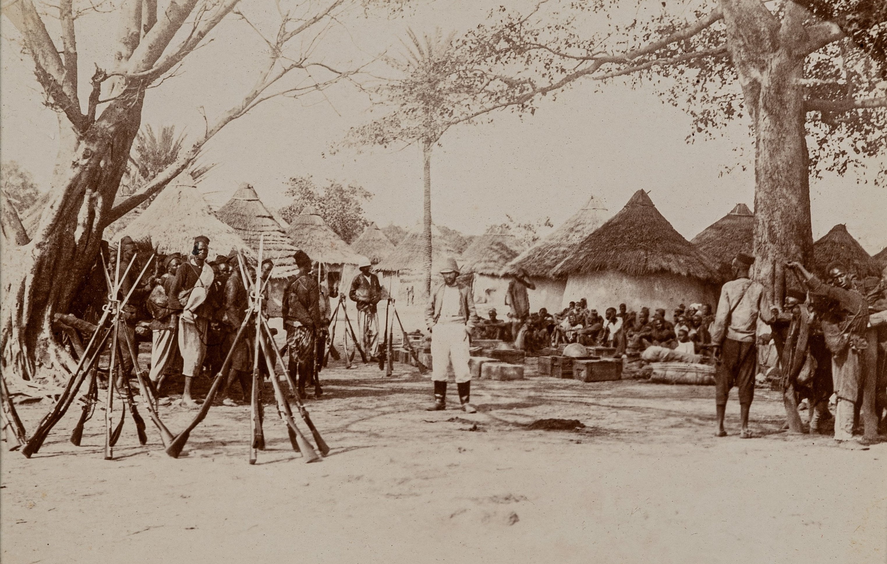 L'image représente une scène historique à l'extérieur d'un village. Au premier plan, plusieurs fusils sont posés verticalement sur le sol, formant une sorte de cadre. Des hommes, en majorité vêtus de vêtements traditionnels, sont rassemblés autour d'une zone centrale. Certains semblent discuter tandis que d'autres restent attentifs, leur posture indiquant un moment de rassemblement communautaire ou de célébration. En arrière-plan, on aperçoit des huttes de forme circulaire, couvertes de toits en paille, typiques des constructions de certaines régions rurales. Plusieurs arbres offrent de l'ombre, ajoutant à l'ambiance naturelle de l'endroit. Le sol est poussiéreux, indiquant le climat aride de la région. Les expressions des visages reflètent un mélange d'intérêt et de sérieux, suggérant une rencontre importante au sein de la communauté.