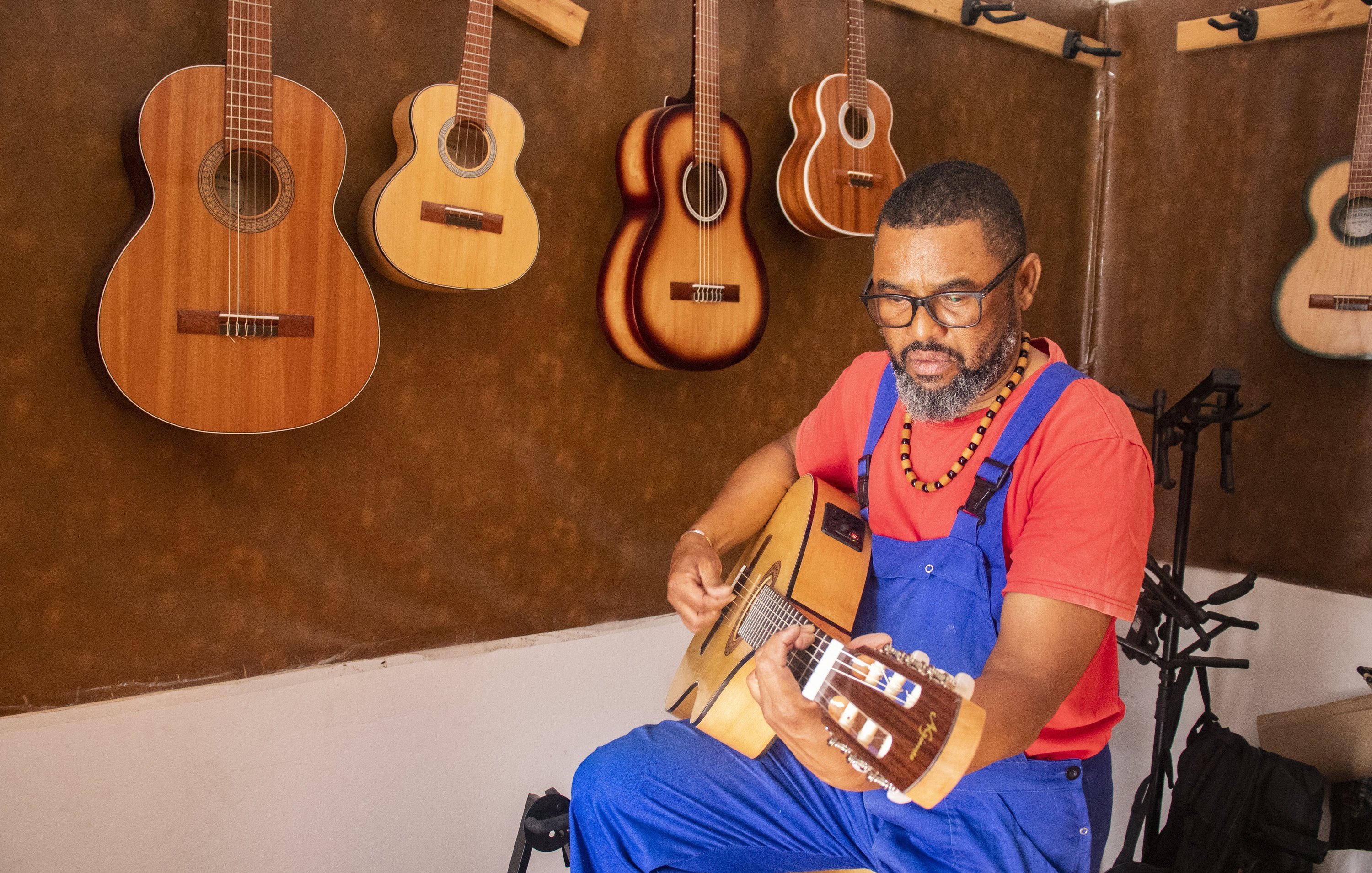 L'image montre un homme assis, vêtu d'une salopette bleue et d'un t-shirt rouge. Il est en train de jouer de la guitare acoustique avec une expression de concentration sur son visage. Autour de lui, plusieurs guitares sont accrochées au mur, dans des teintes de bois naturel. L'ambiance de l'espace est chaleureuse et artistique, soulignant sa passion pour la musique.