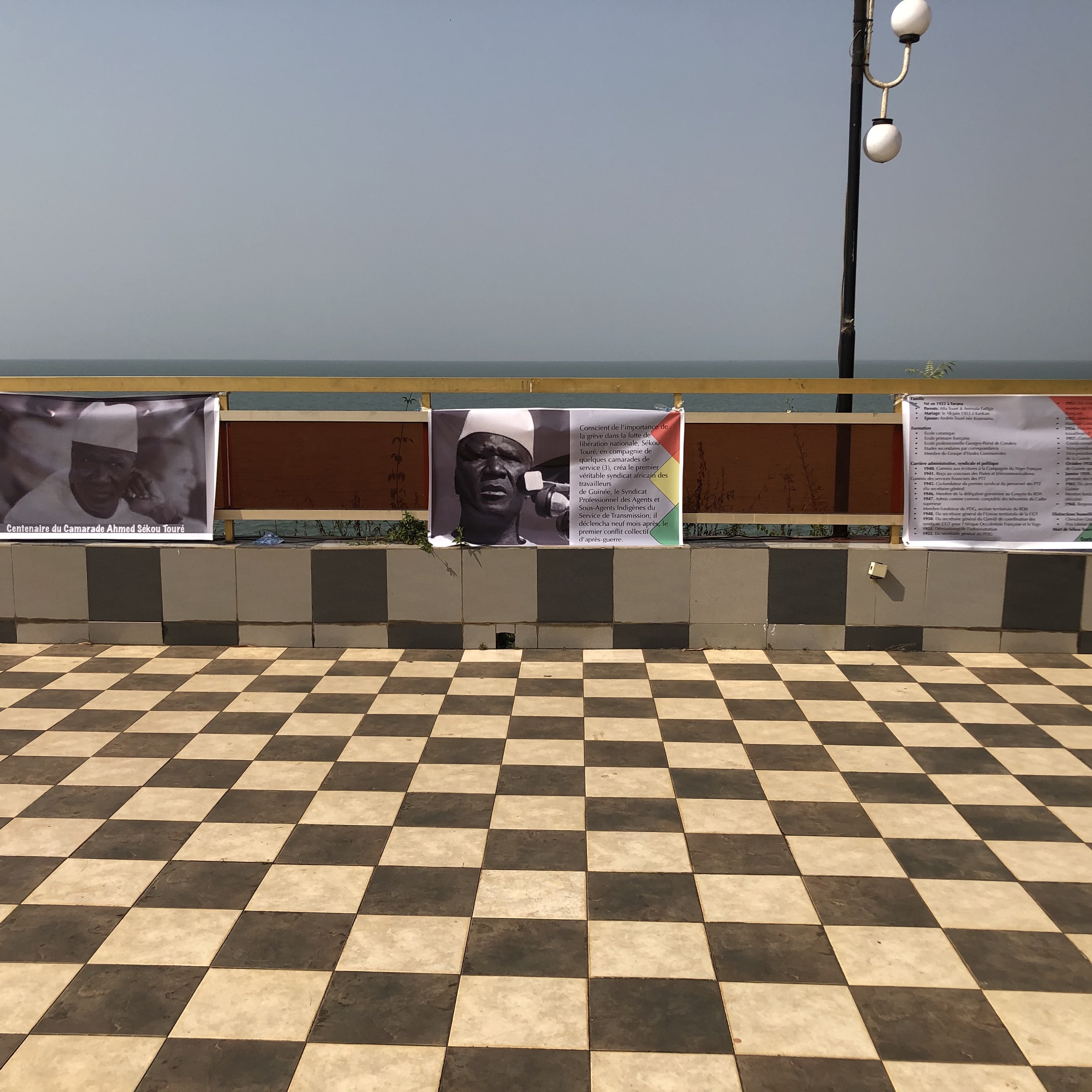 L'image montre un espace extérieur avec un sol en carreaux de damier, alternant entre des teintes claires et foncées. En arrière-plan, on aperçoit une étendue d'eau qui pourrait être la mer ou un lac, sous un ciel clair. Sur un muret, plusieurs affiches sont accrochées. Ces affiches semblent contenir du texte et des images en rapport avec un événement ou une exposition. L'environnement dégage une atmosphère calme, avec un éclairage naturel et une vue agréable sur l'eau.