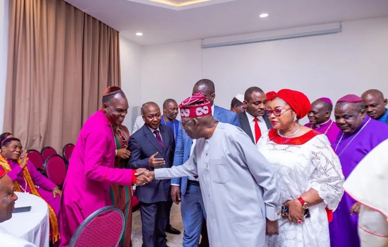 L'image montre un groupe de personnes rassemblées dans une salle, probablement lors d'un événement formel. Au centre, un homme en tenue traditionnelle serre la main d'un autre homme, qui porte une chemise claire. Les autres participants, habillés de manière variée, se tiennent autour d'eux. Certaines femmes portent des vêtements colorés et des accessoires élégants. L'ambiance semble conviviale et respectueuse, avec des sourires et des gestes amicaux. Les murs de la salle sont de couleur claire, et il y a des chaises rouges disposées autour de tables.