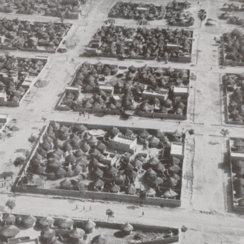 L'image montre une vue aérienne d'un village africain. On peut voir un agencement organisé des habitations, qui sont principalement des maisons traditionnelles en toit de chaume, caractéristiques de certaines régions. Les structures sont regroupées en plusieurs blocs délimités par des chemins. Des arbres sont dispersés dans l'espace, ajoutant une touche de verdure. Quelques personnes se déplacent le long des routes, suggérant une animation dans ce décor rural. Le photographique est en noir et blanc, ce qui donne une atmosphère historique à la scène.